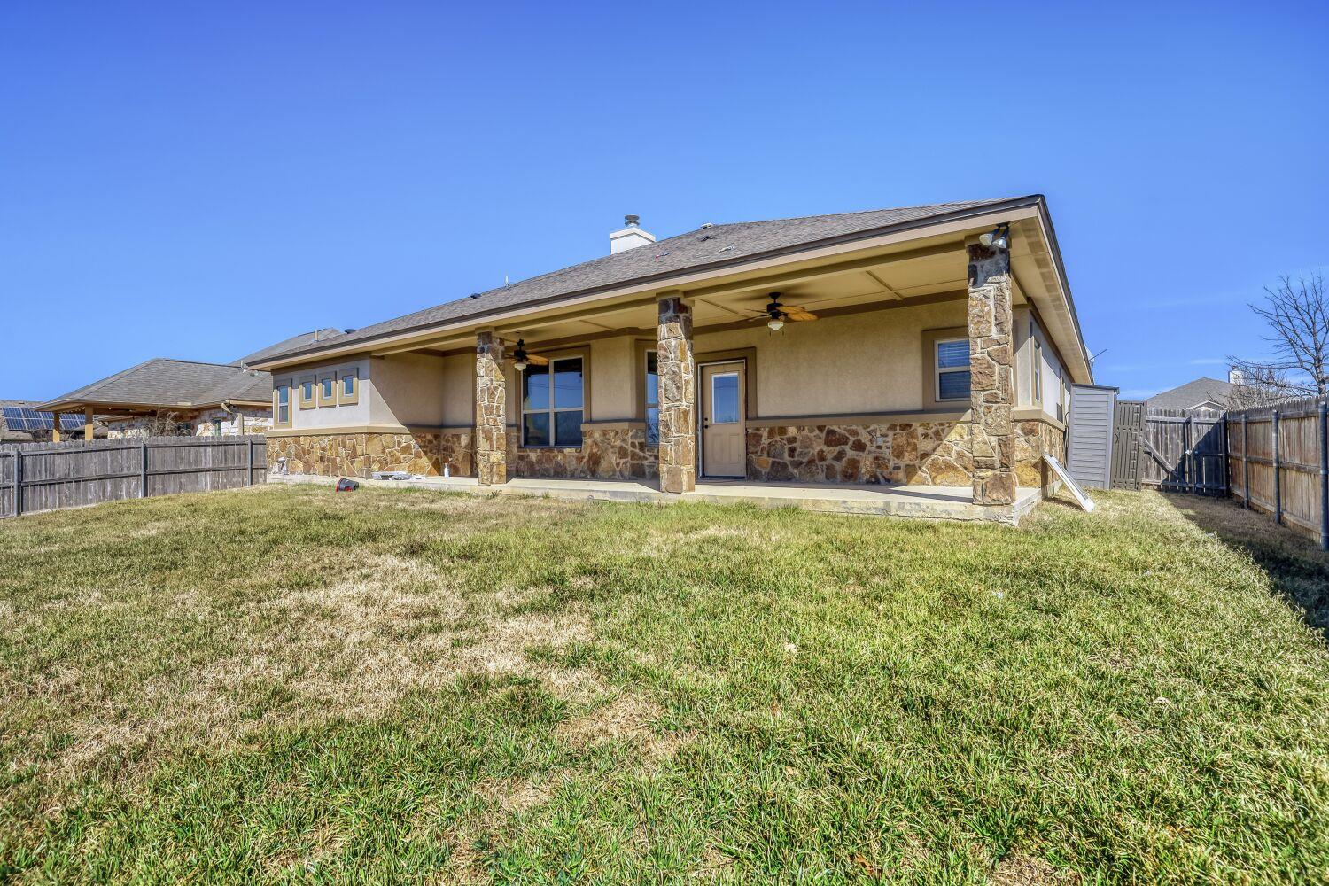 Rear of a single-story home with a covered stone-column patio and fenced grassy backyard.