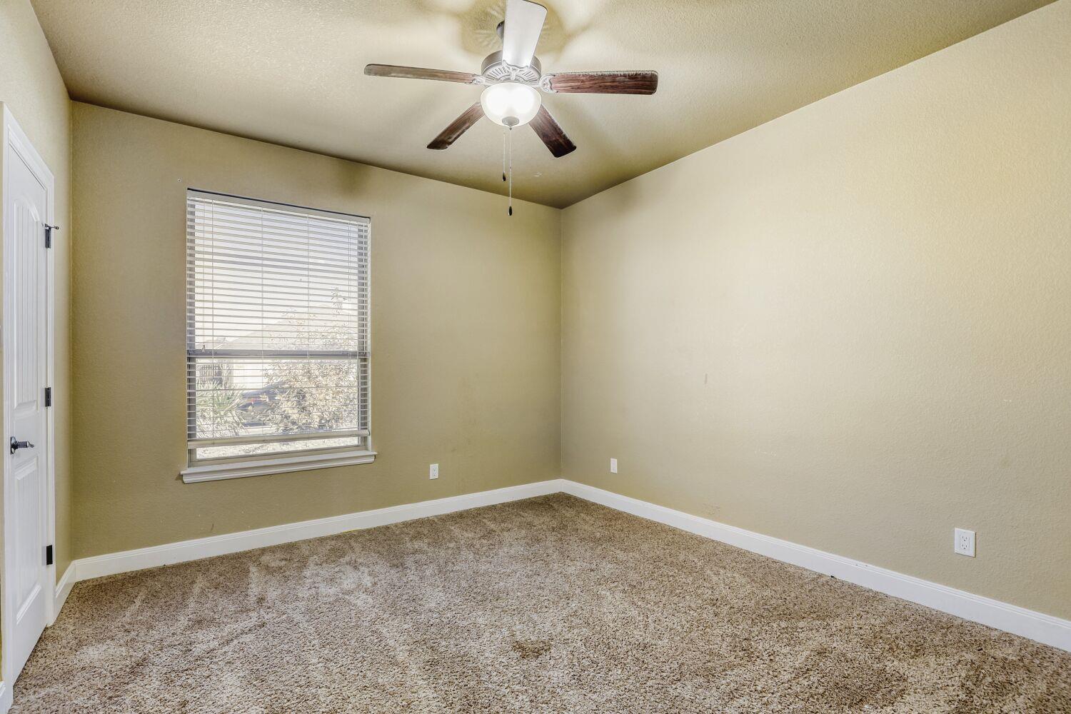 Empty bedroom with patterned carpet, window blinds, and a ceiling fan with light.