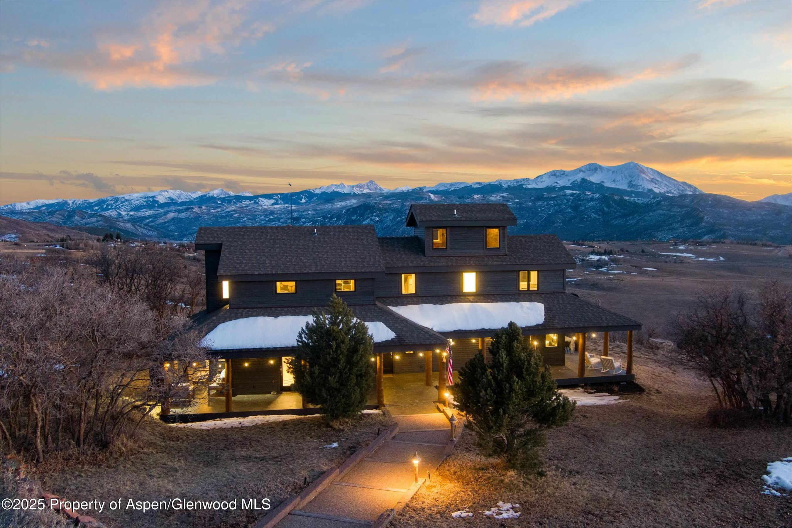 Dark mountain home with a lit wraparound porch and snow-covered peaks at sunset.