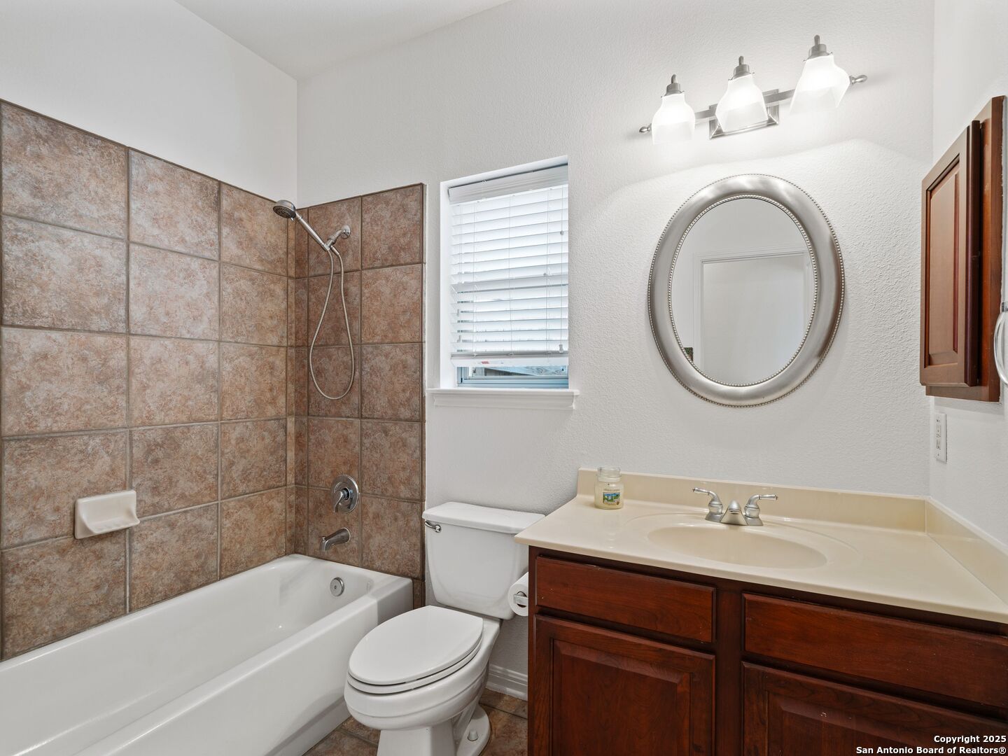 Bathroom with a tan tiled tub and shower, dark wood vanity, oval mirror, and window with blinds.