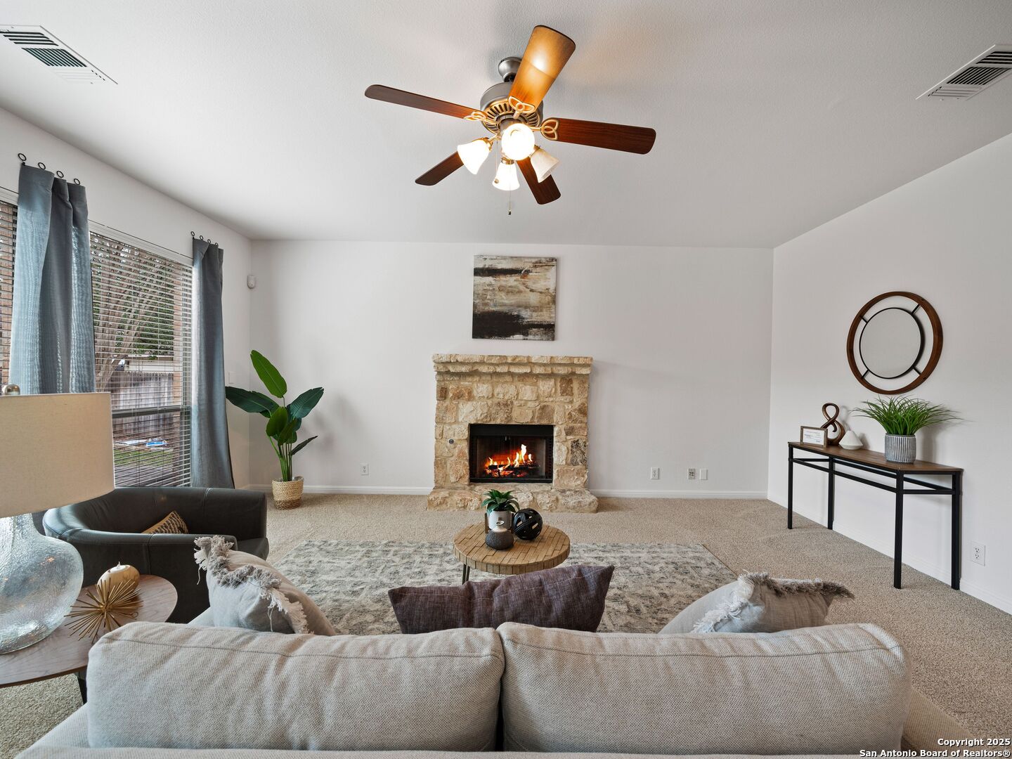 Living room with a stone fireplace, lit fire, ceiling fan, and neutral furnishings.