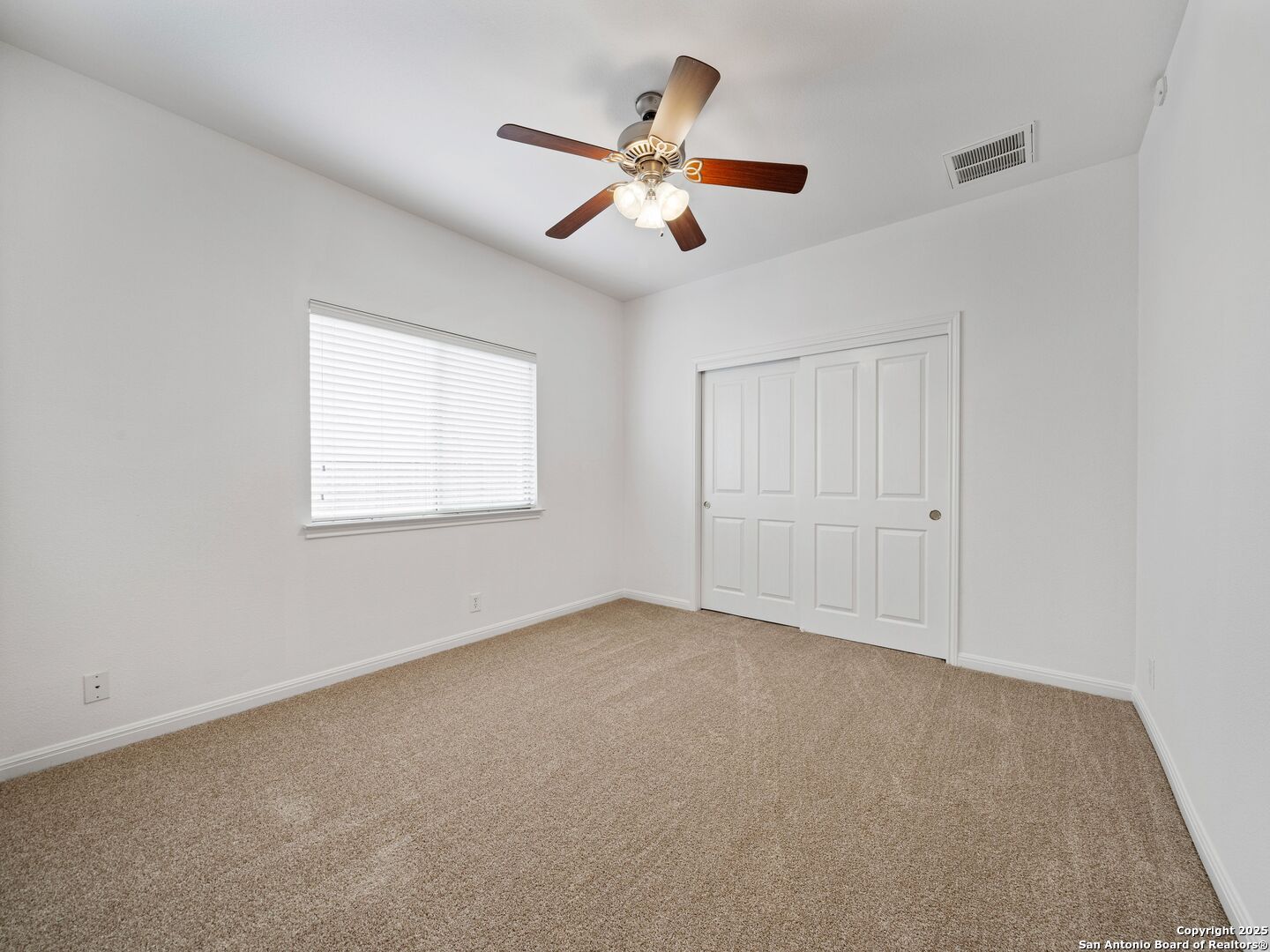 Empty bedroom with beige carpet, double closet doors, and a ceiling fan with lights.