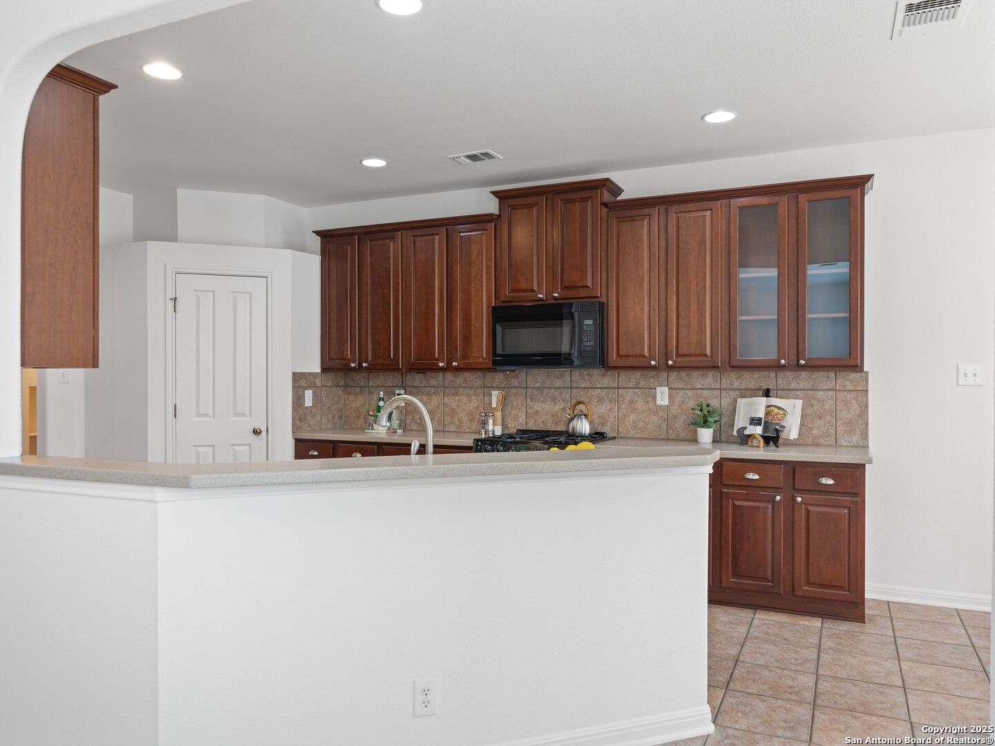 Kitchen with dark wood cabinetry, glass-front upper cabinets, tiled backsplash, and a broad peninsula.