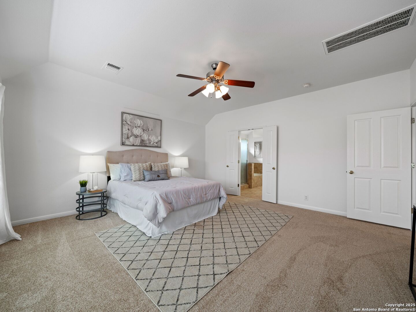 Staged bedroom with a large bed, diamond-pattern rug, and doorway to an adjoining bathroom.
