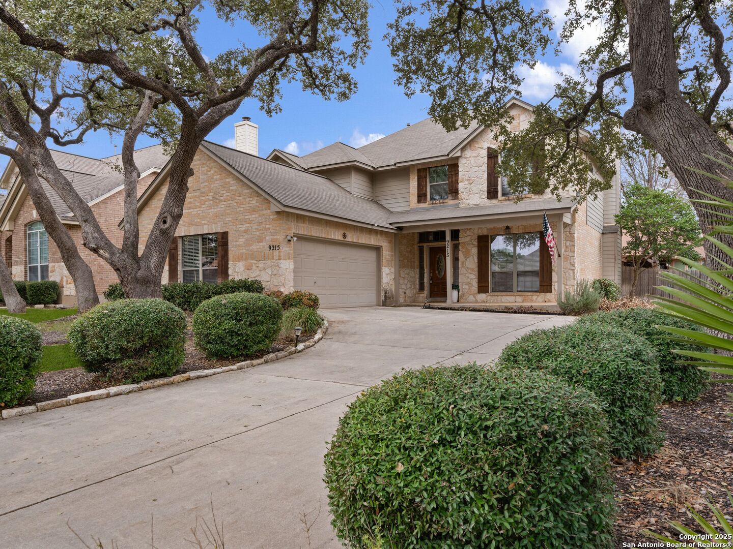 Two-story stone and brick house with a garage, broad driveway, and mature oak trees.