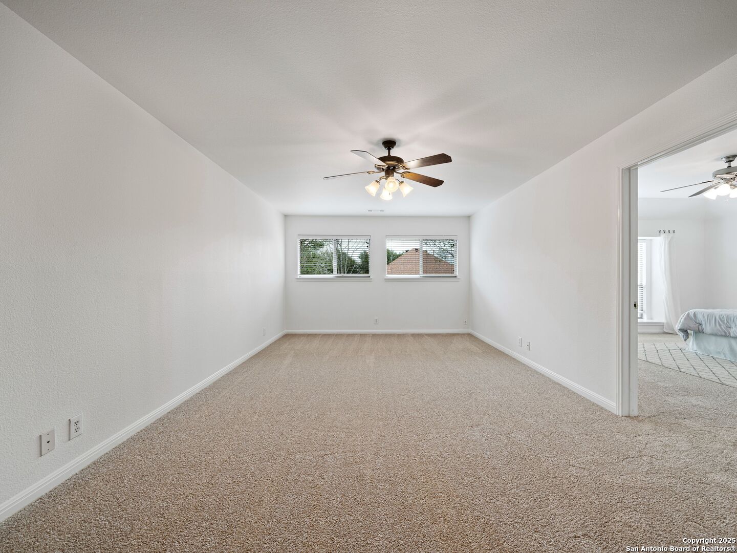 Spacious carpeted room with a ceiling fan, two windows, and a doorway to a bedroom.