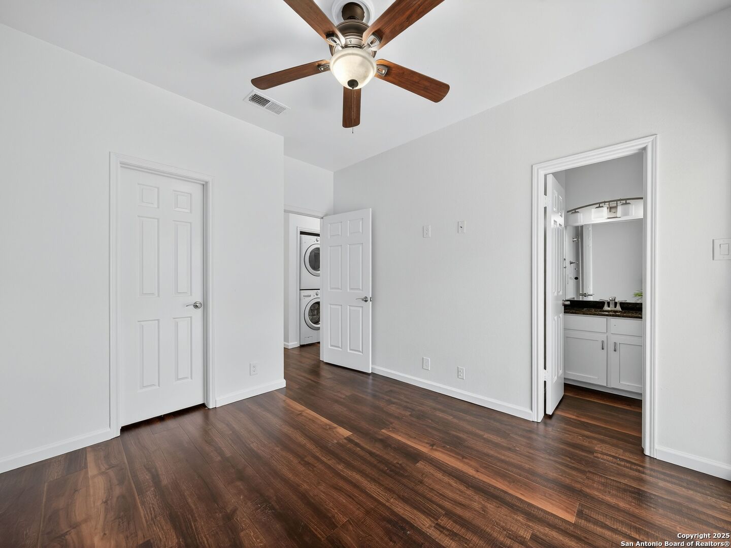Empty bedroom with dark wood floors, a ceiling fan, and doorways to the bathroom and laundry area.