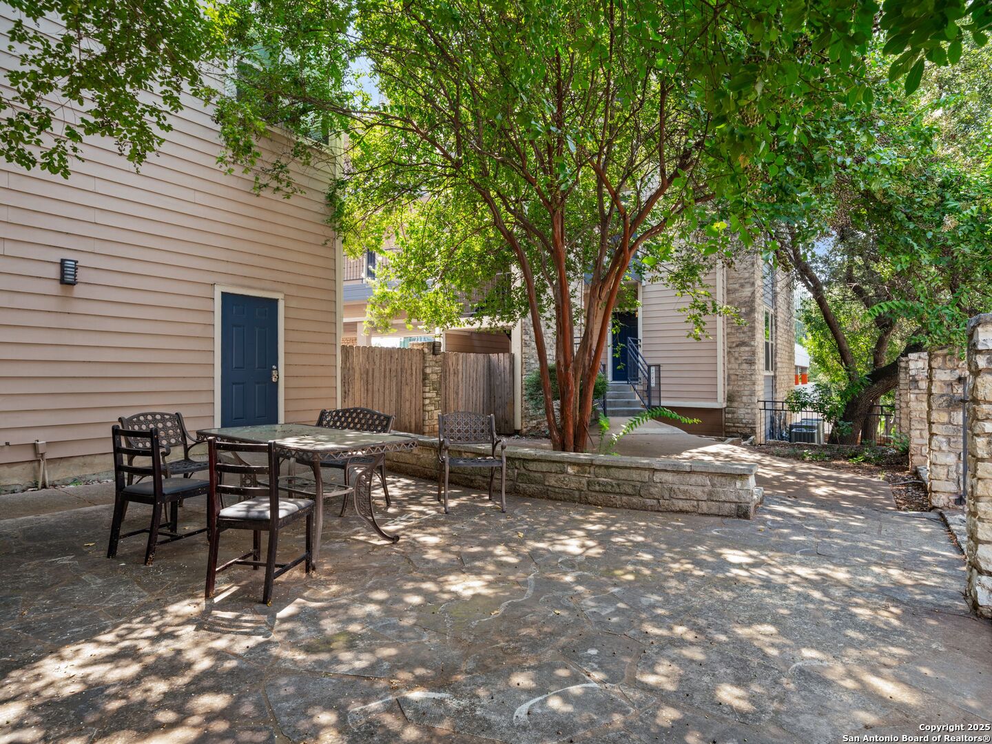 Apartment courtyard with a stone patio, outdoor dining set, and mature shade trees.