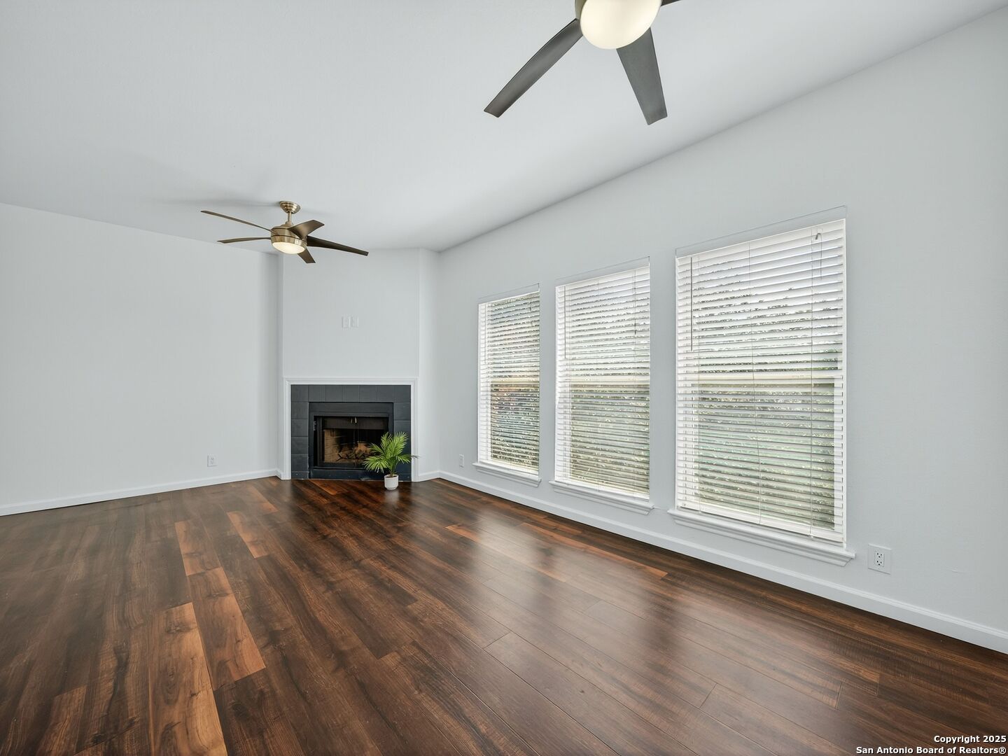 Empty living room with hardwood floors, a fireplace, three windows, and two ceiling fans.
