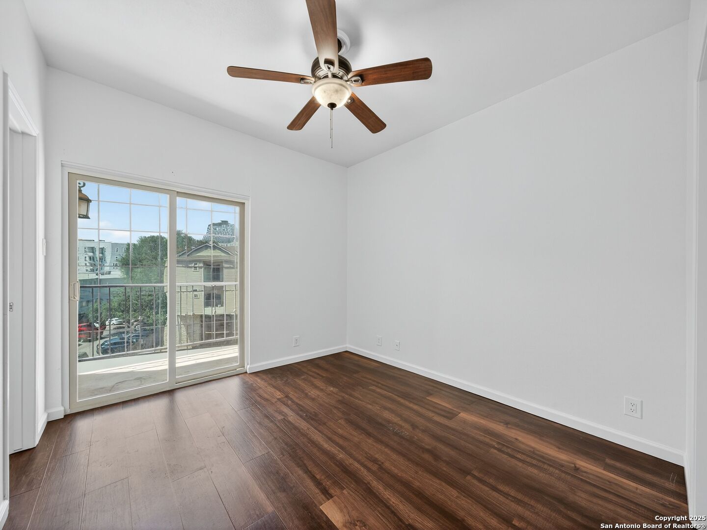 Empty room with dark wood flooring, ceiling fan, and sliding glass door to a balcony.
