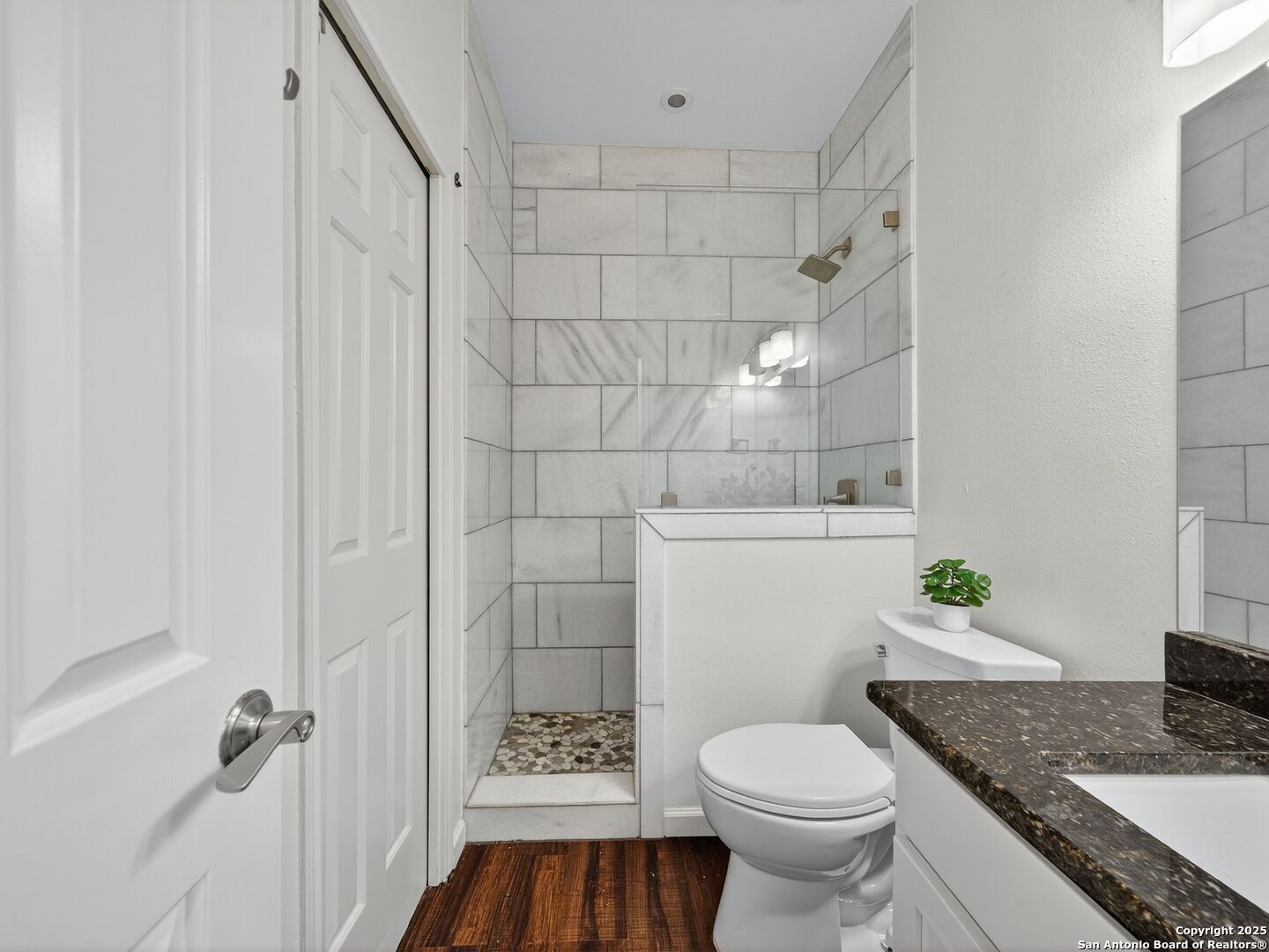 Bathroom with a marble-look tiled shower, pebble floor, white toilet, and dark stone vanity countertop.