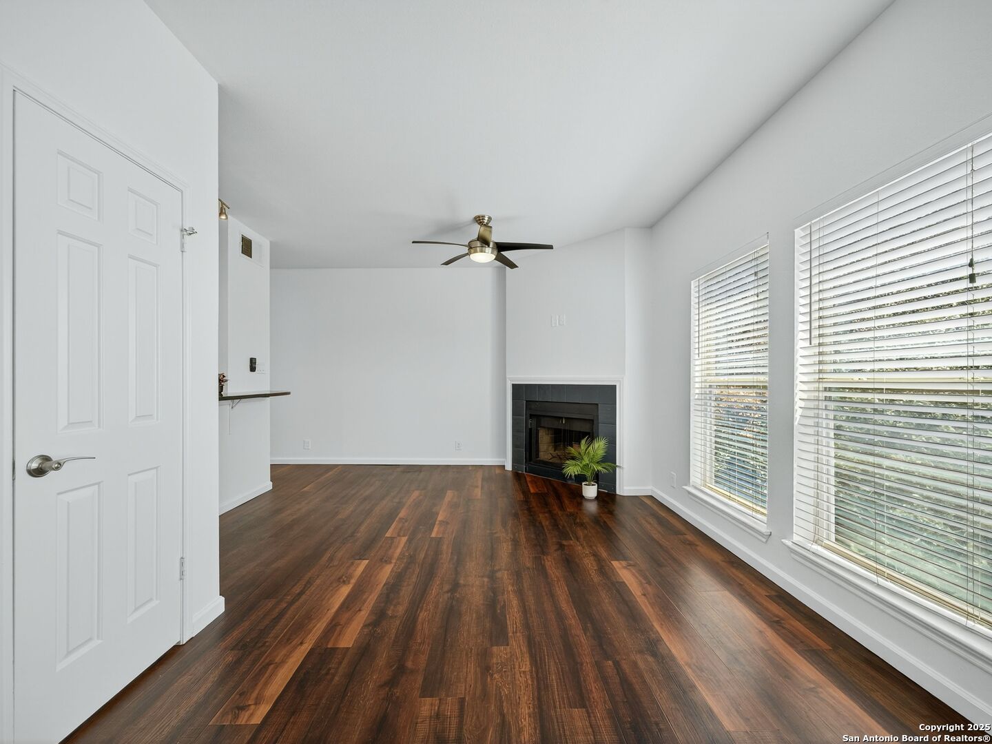 Vacant living room with dark wood floors, a corner fireplace, and large windows with blinds.