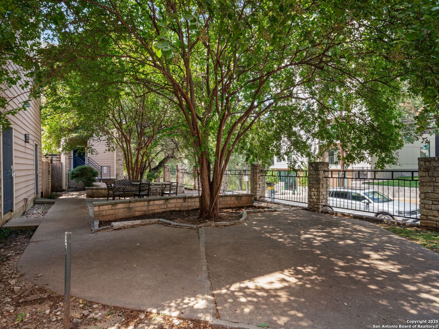 Shaded apartment courtyard with mature trees, patio seating, stone walls, and a metal gate.