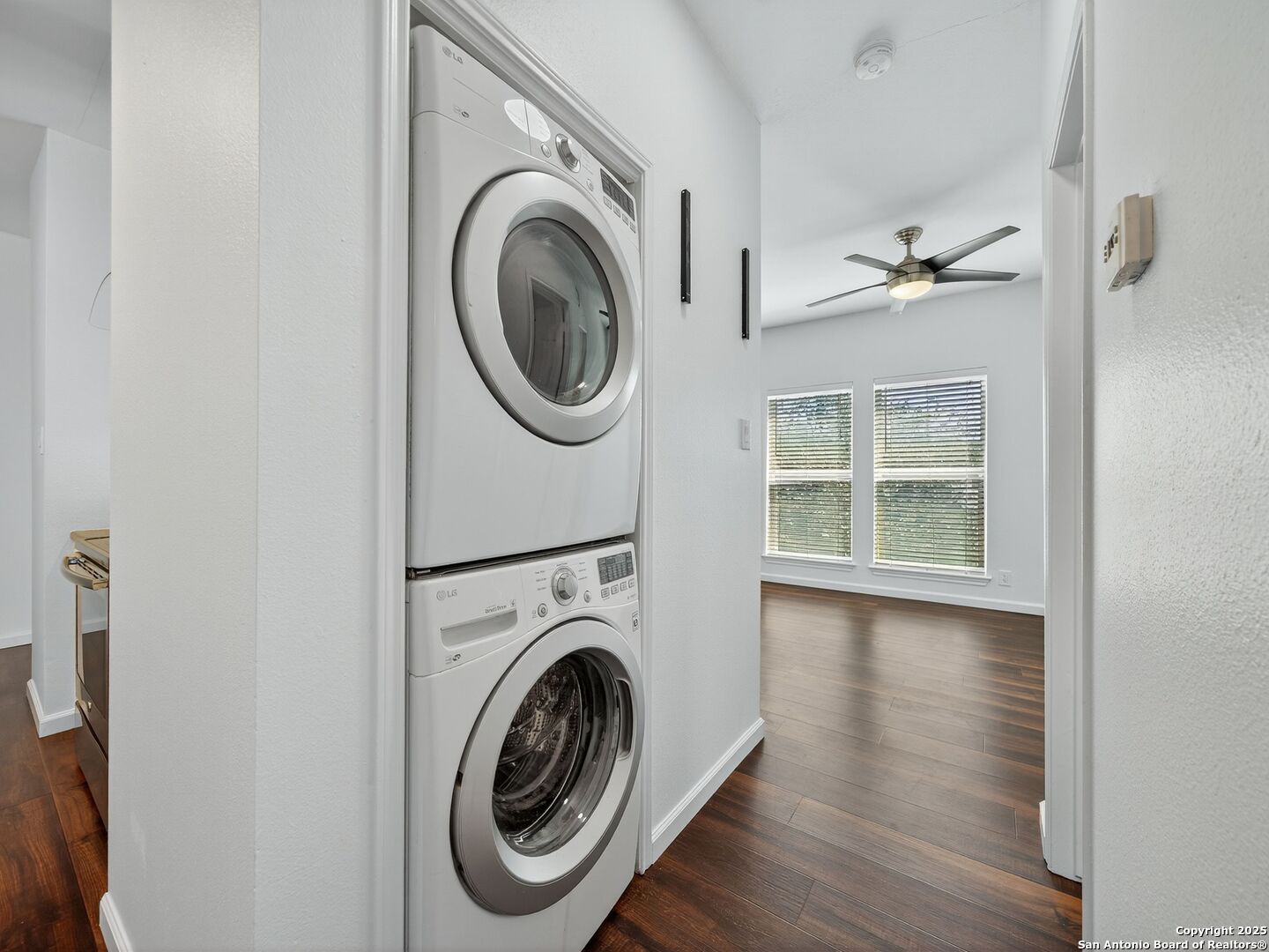 Stacked washer and dryer beside an unfurnished room with wood flooring and two windows.