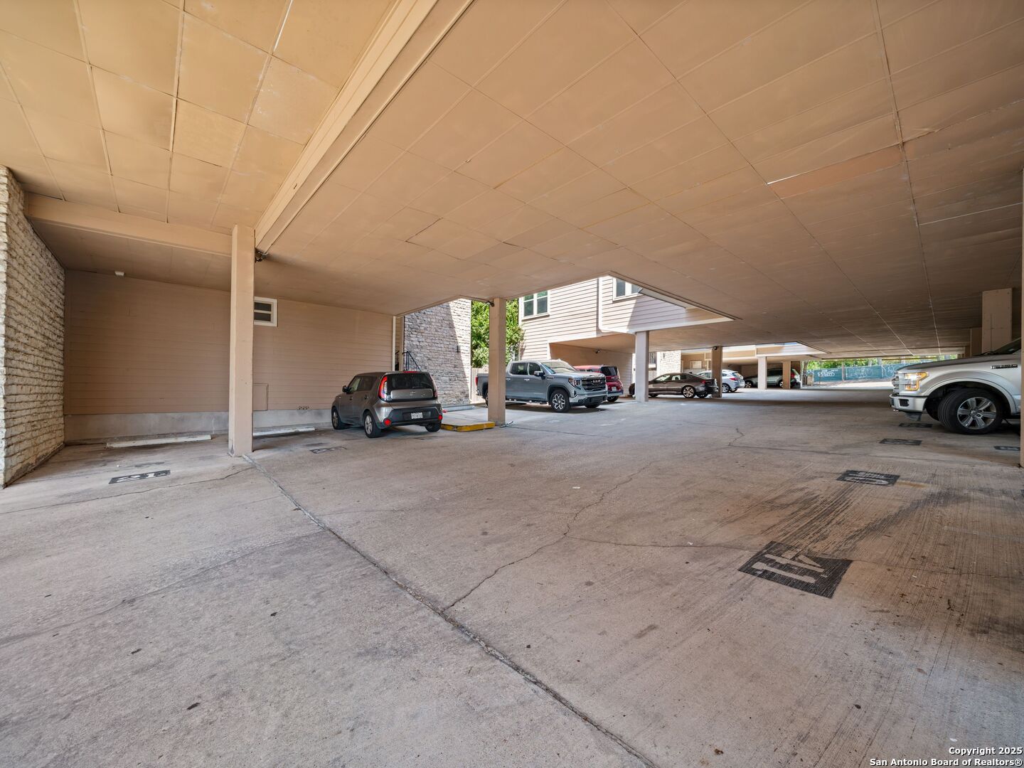 Covered apartment parking area with concrete paving and several parked vehicles.