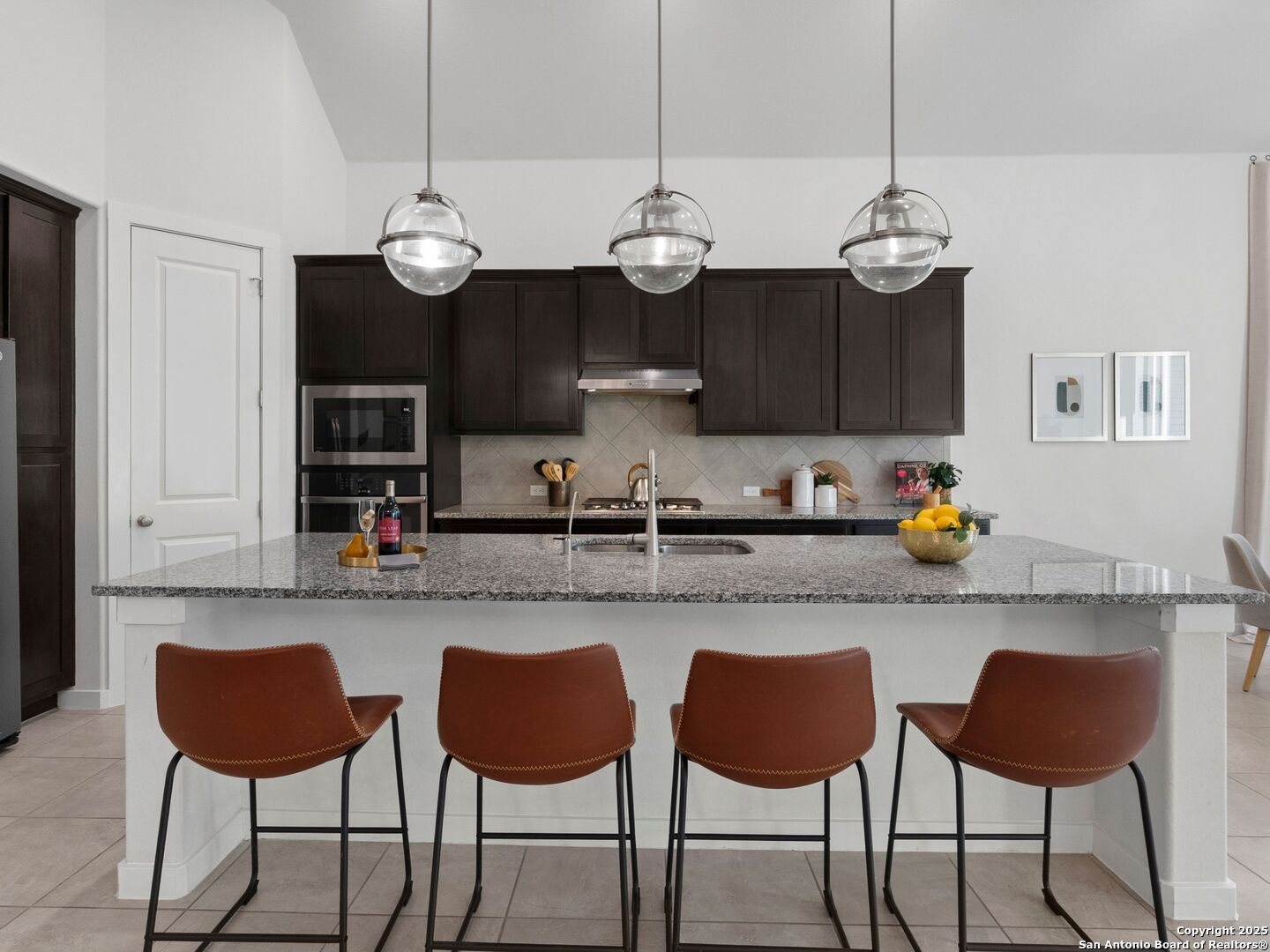 Kitchen with dark cabinets, a large stone island, four brown bar stools, and glass pendant lights.