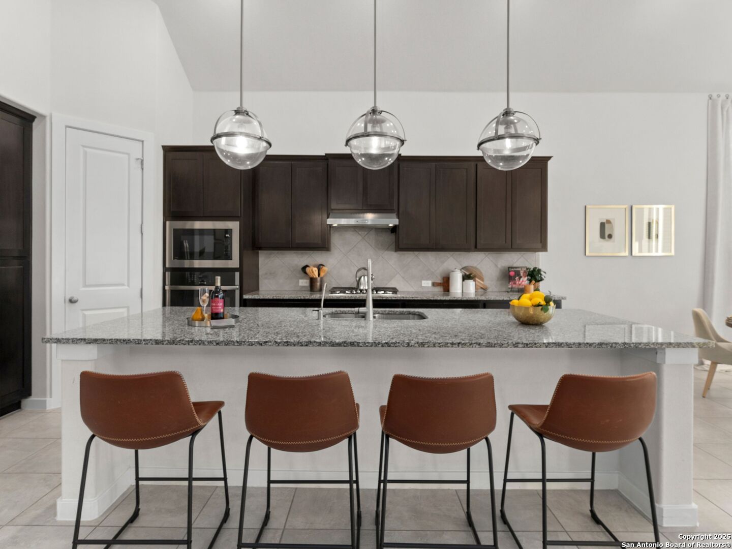 Kitchen with dark wood cabinets, granite island, four brown bar stools, and glass pendant lights.