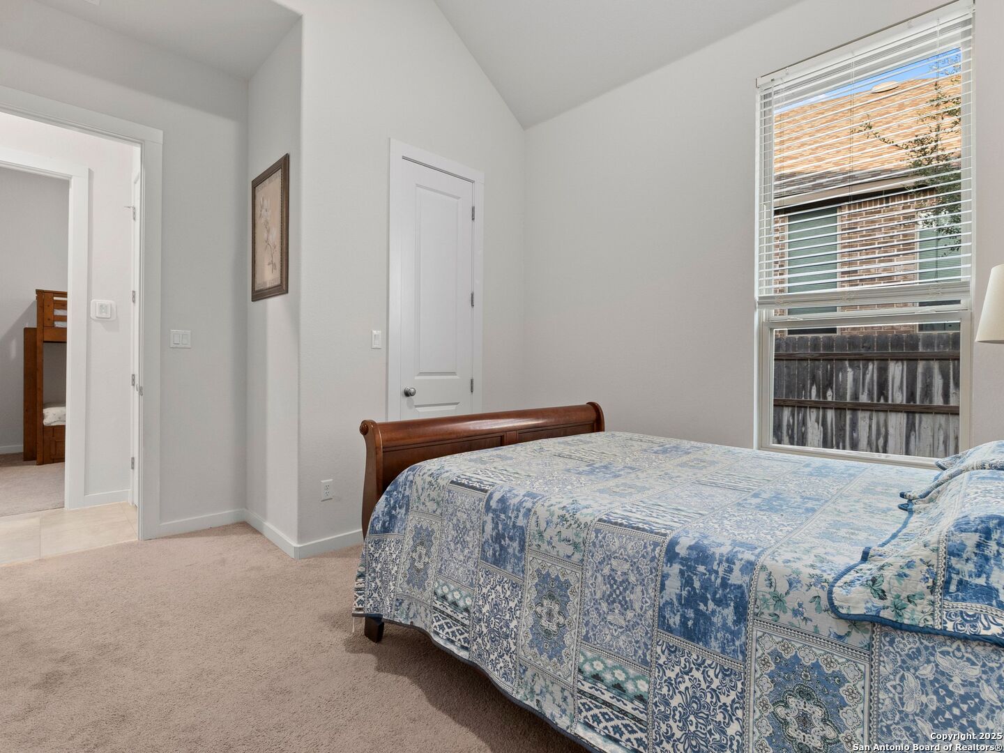 Bedroom with a wooden bed, blue-and-white patterned quilt, beige carpet, and a large window with blinds.