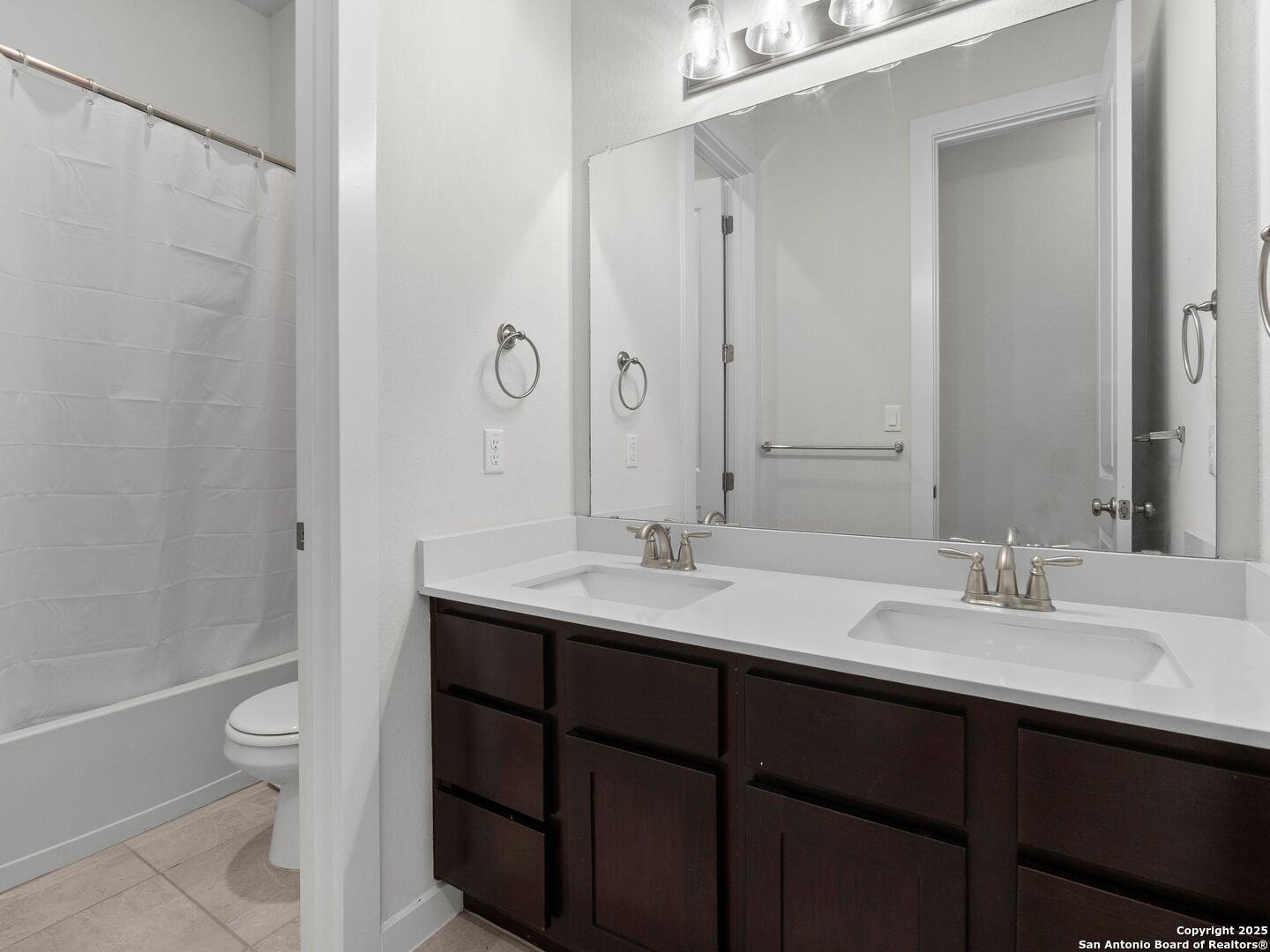 Bathroom with a dark double-sink vanity and large mirror, with the shower and toilet visible behind a partial wall.