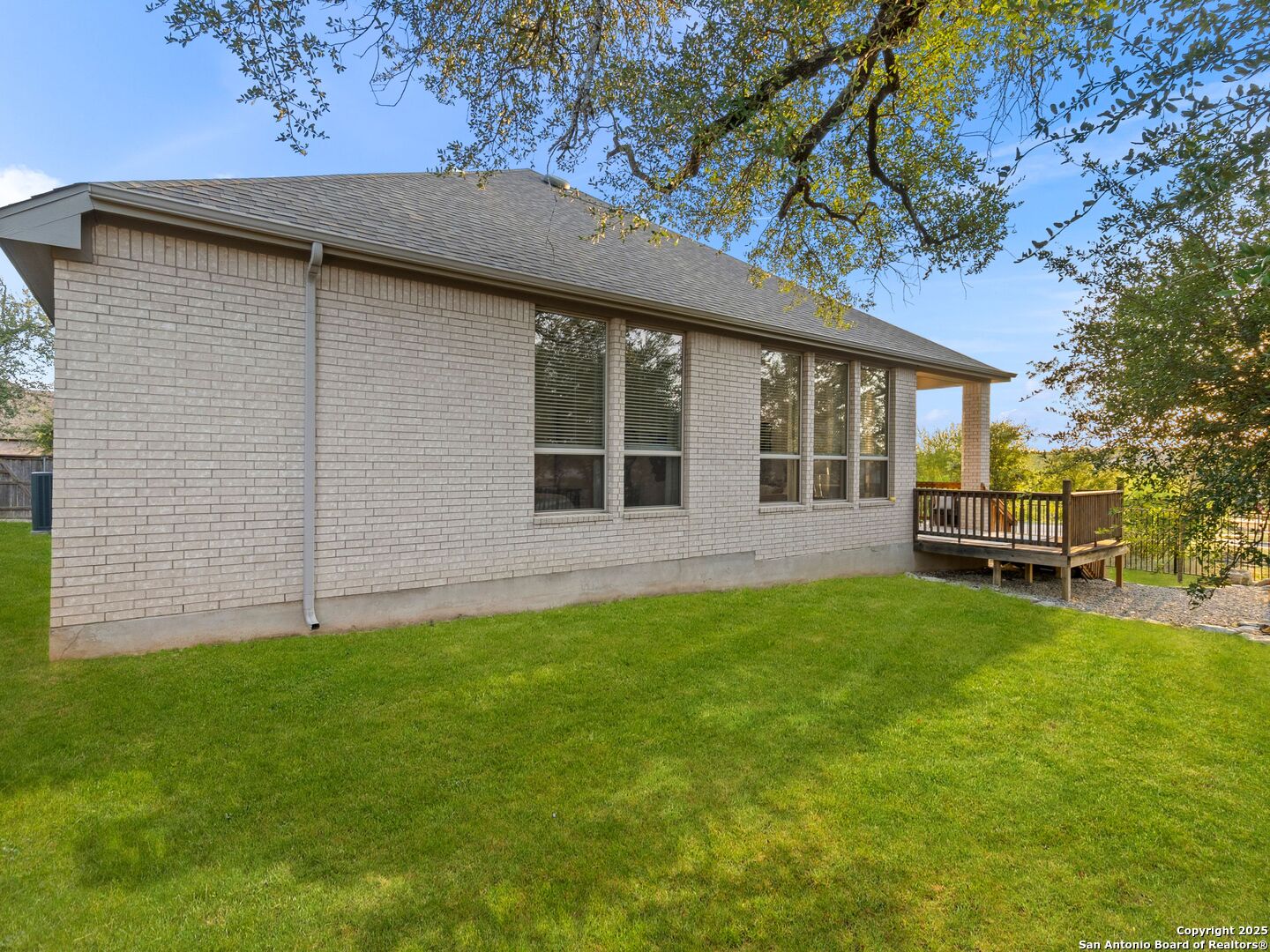 Light brick house with a small wooden deck overlooking a broad green lawn beneath mature trees.