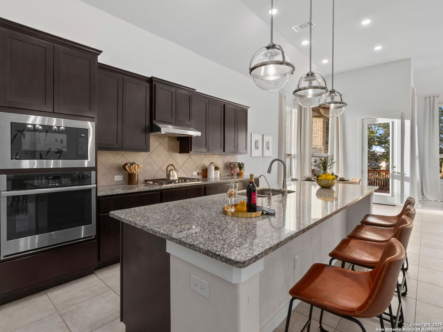 Kitchen with dark cabinetry, double ovens, and a large stone island with four leather bar stools.