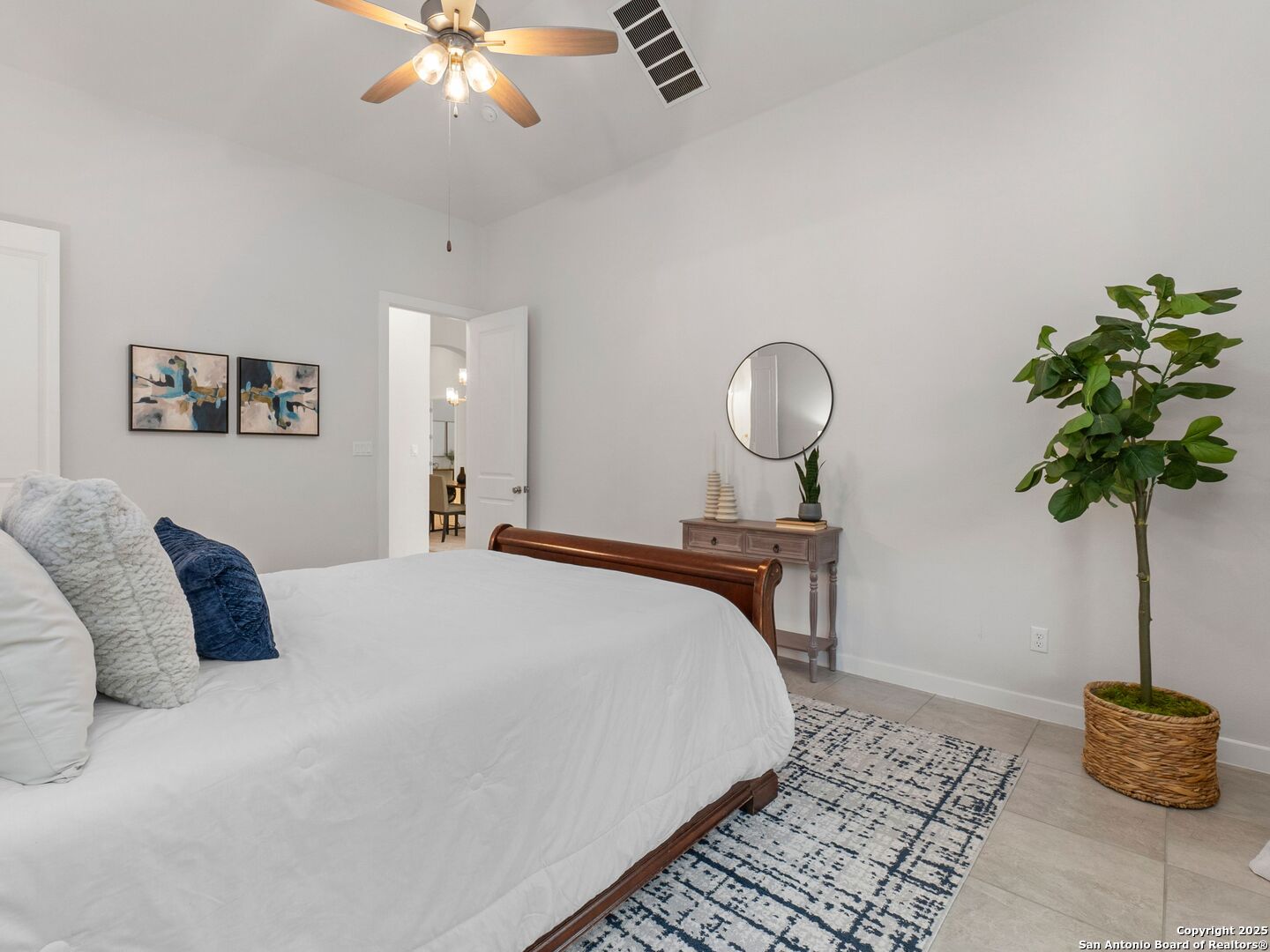 Furnished bedroom with a white bed, patterned rug, ceiling fan, and potted tree.