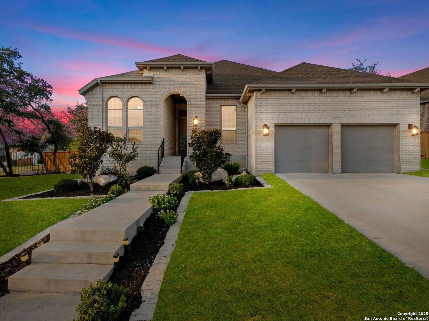 Light brick home with an arched entry, two-car garage, and landscaped front lawn at dusk.