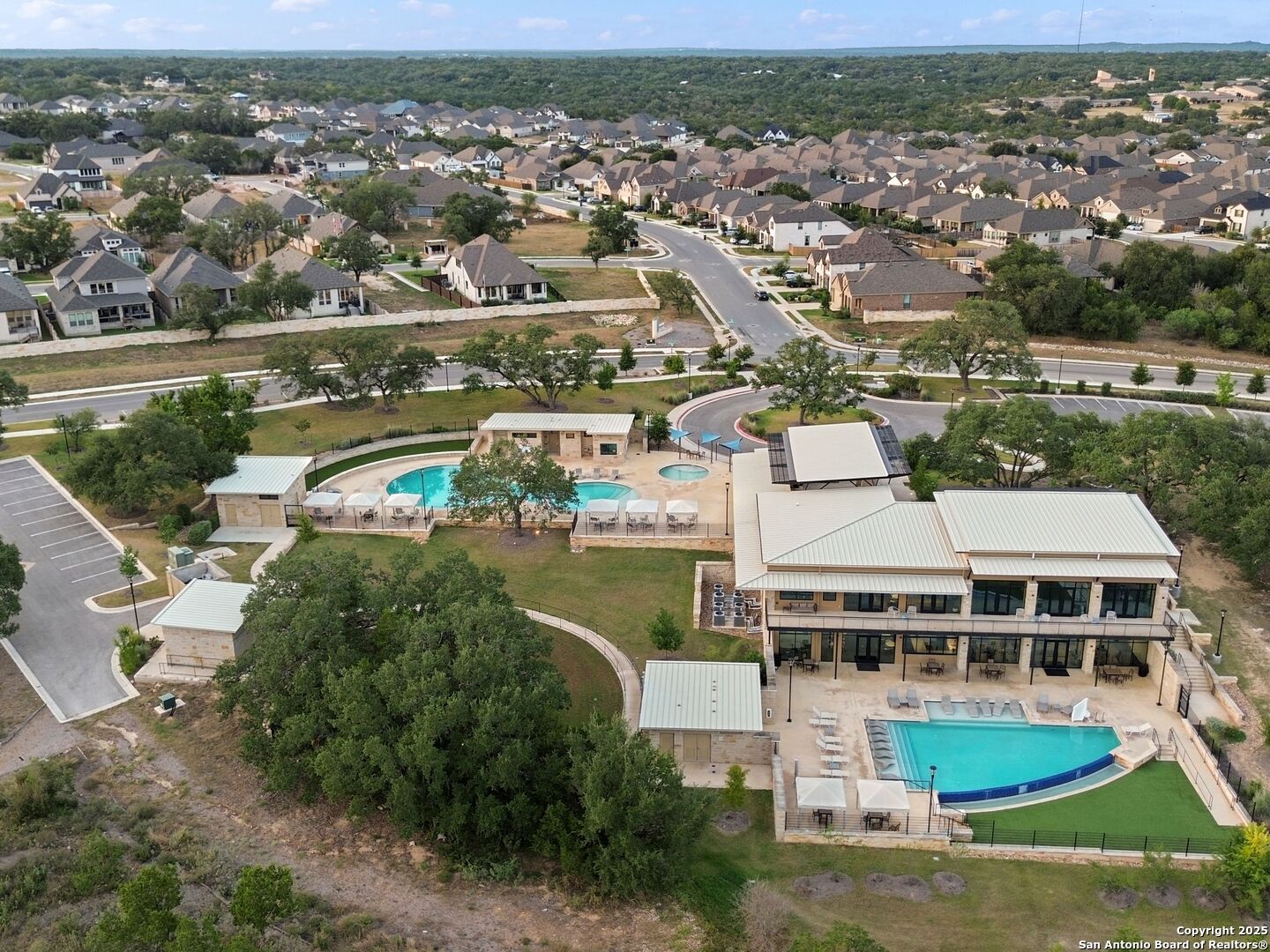 Aerial view of a neighborhood clubhouse and two swimming pools, with homes and wooded land beyond.