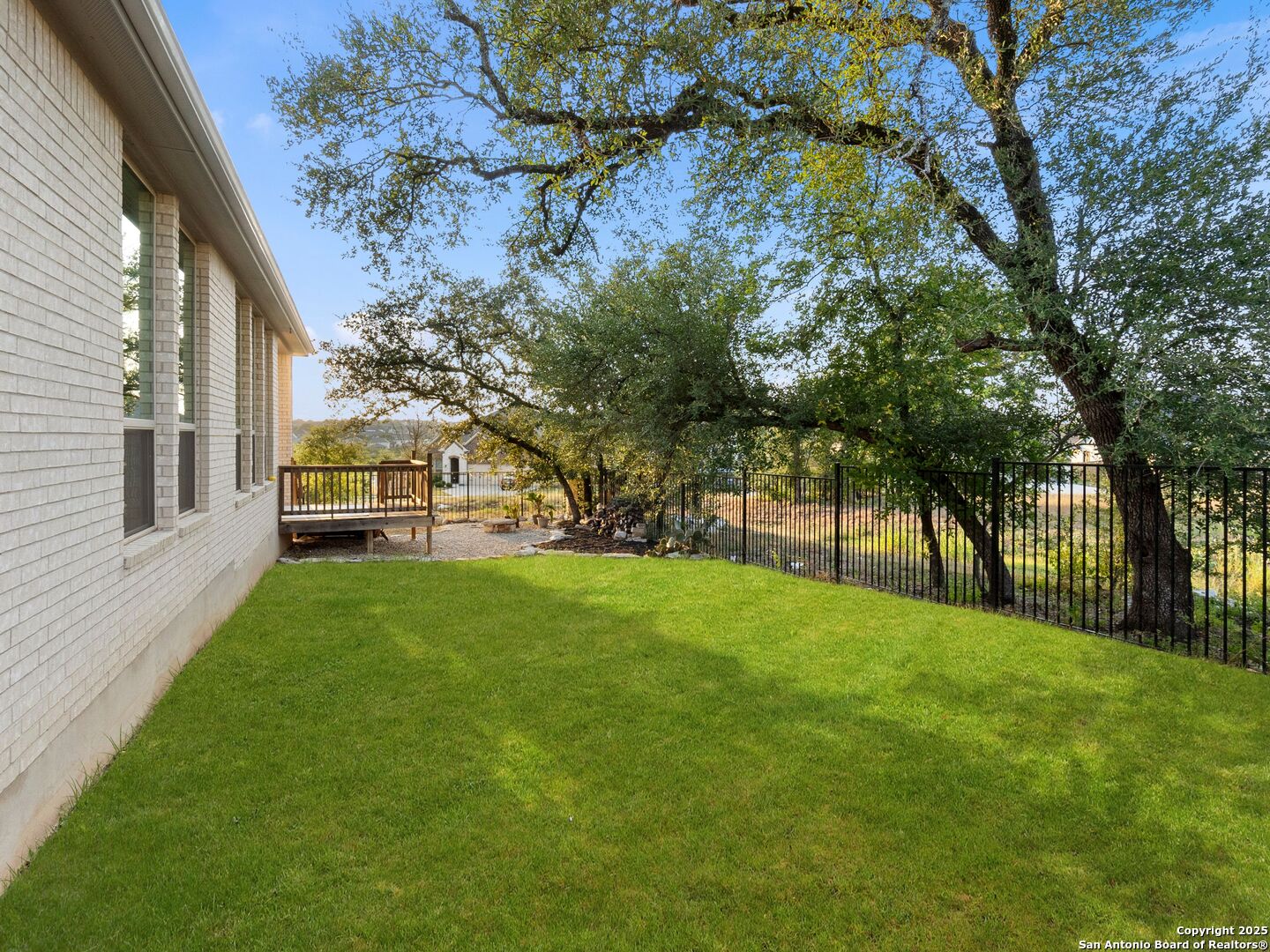 Brick home’s side yard with a broad lawn, small deck, mature oak trees, and black metal fence.