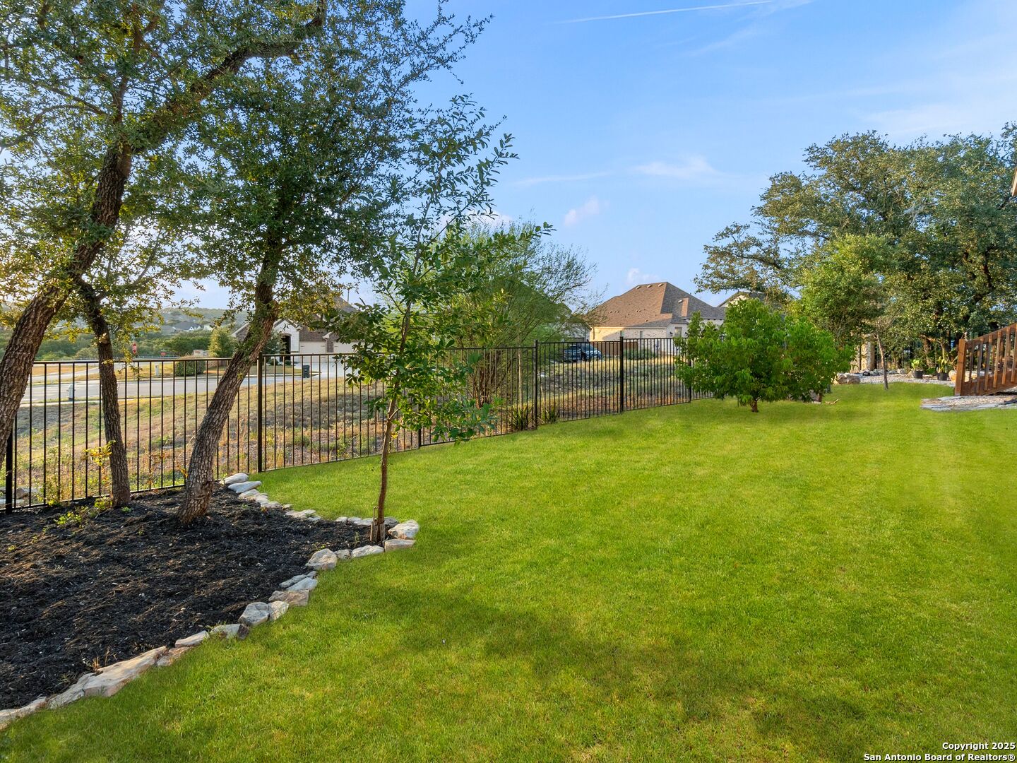 Backyard lawn bordered by mature trees, landscaped beds, and a black metal fence.