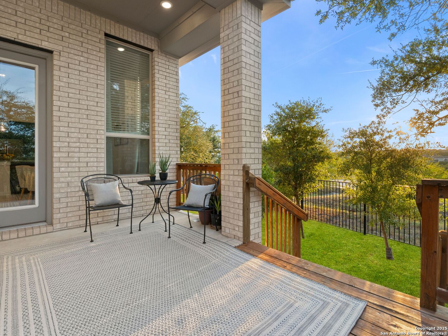 Covered brick patio with seating and a wooden deck overlooking a fenced, tree-lined yard.