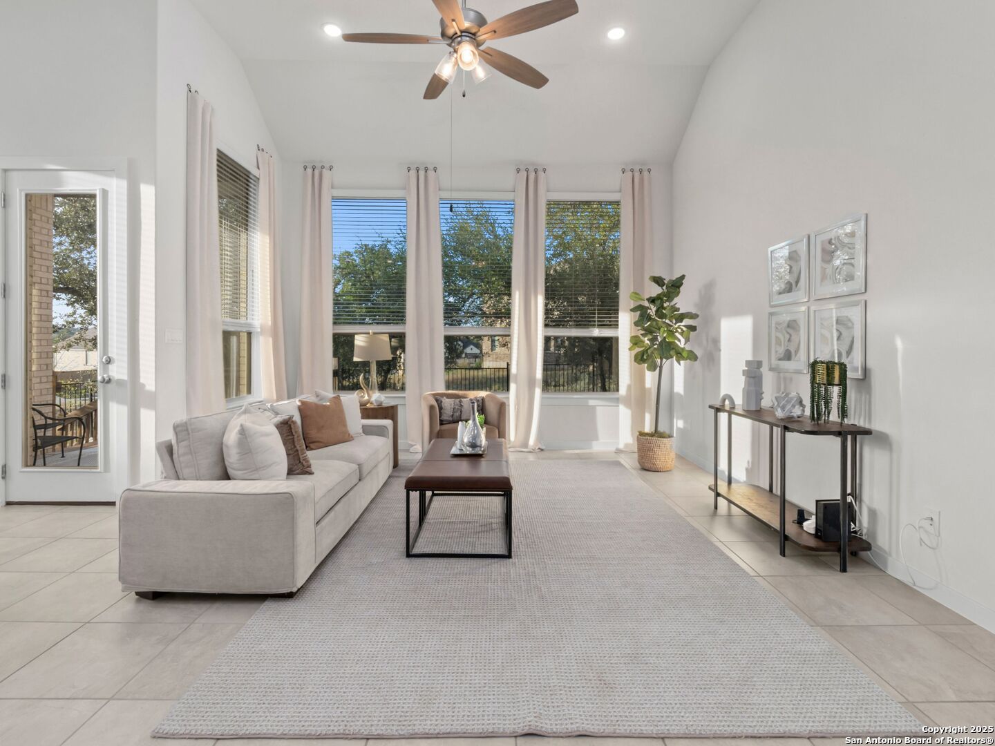 Staged living room with vaulted ceiling, large windows, gray sofa, and pale area rug.