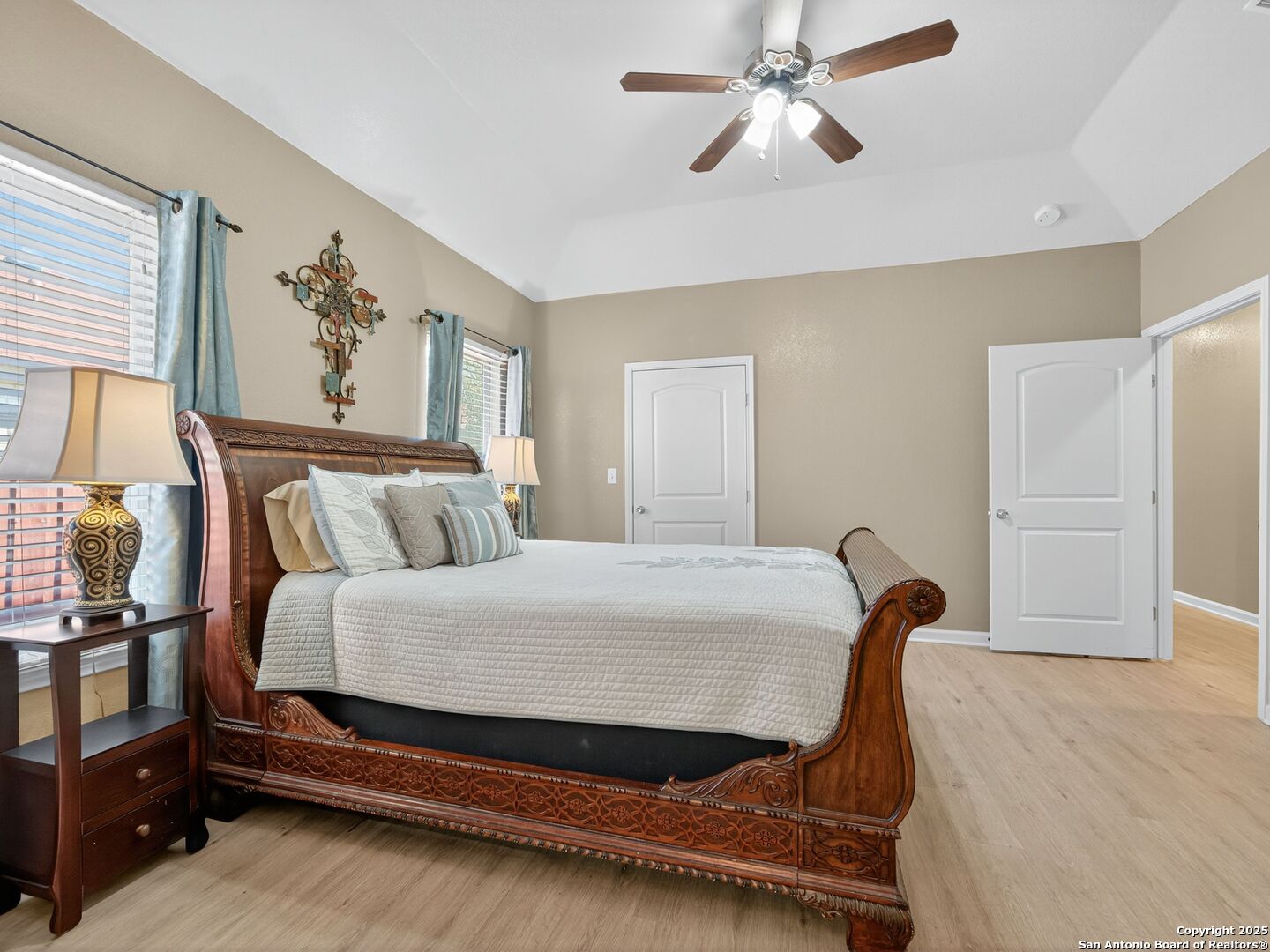 Furnished bedroom with a carved wood bed, pale quilt, blue curtains, and ceiling fan.