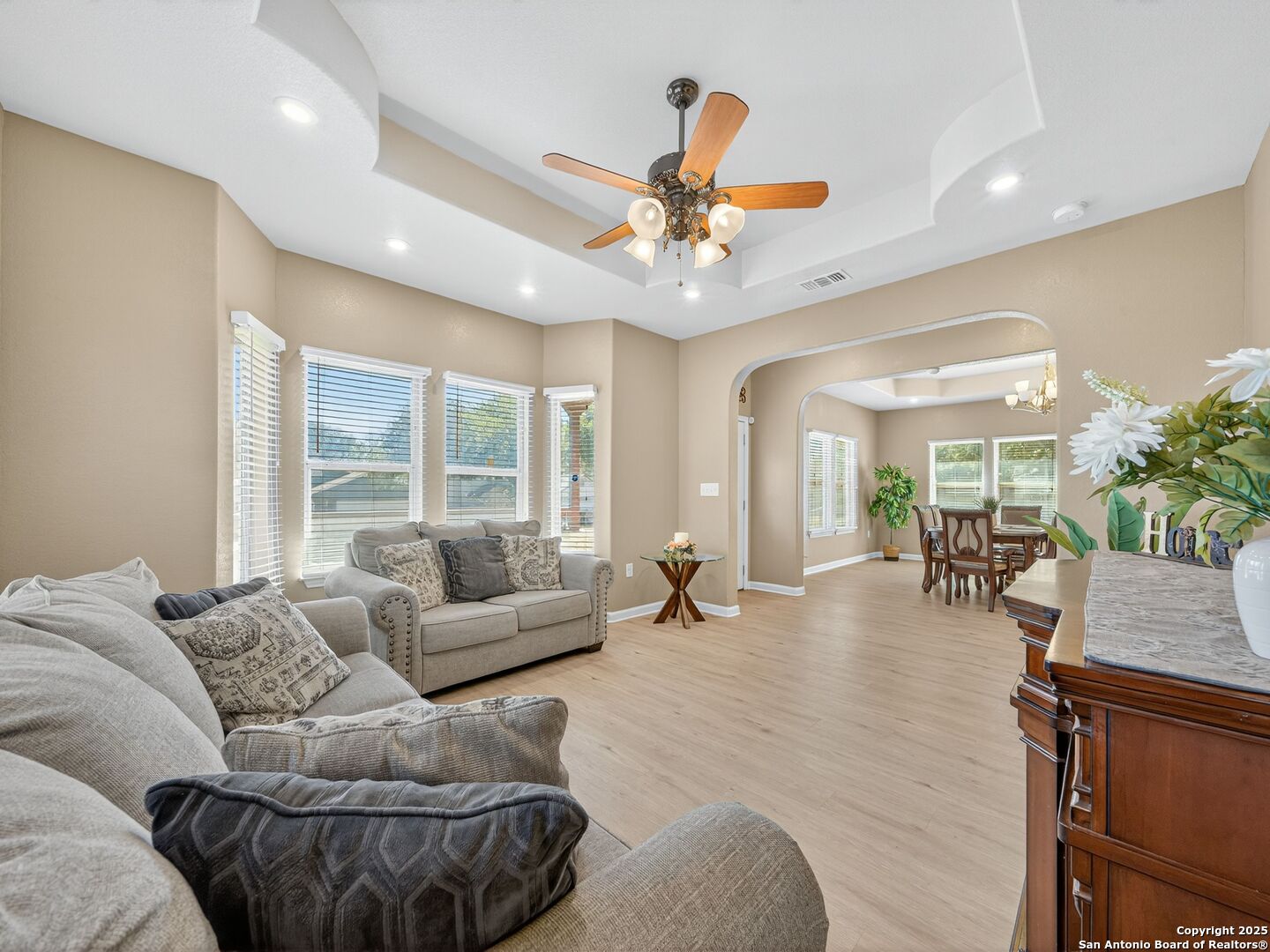 Living room with beige sofas and a ceiling fan, opening to a dining area.