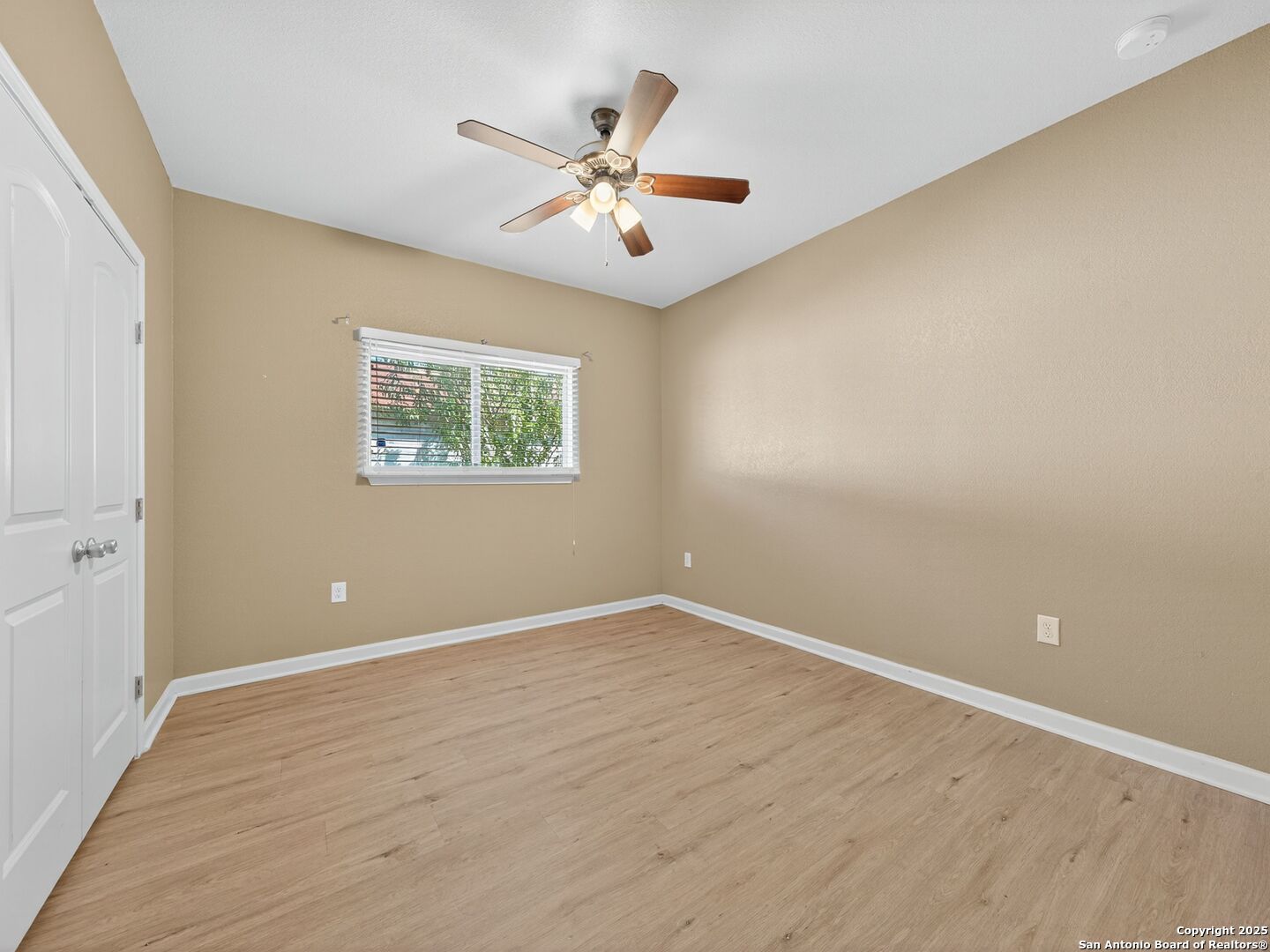 Empty bedroom with light wood-look flooring, a window with blinds, closet doors, and a ceiling fan.