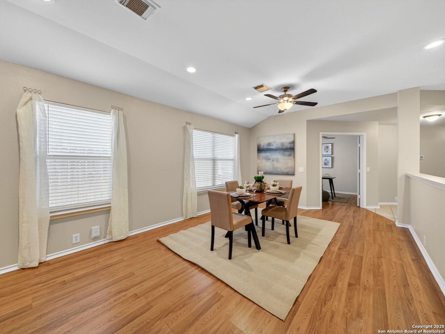 Dining area with a set table, two curtained windows, warm wood flooring, and a ceiling fan.