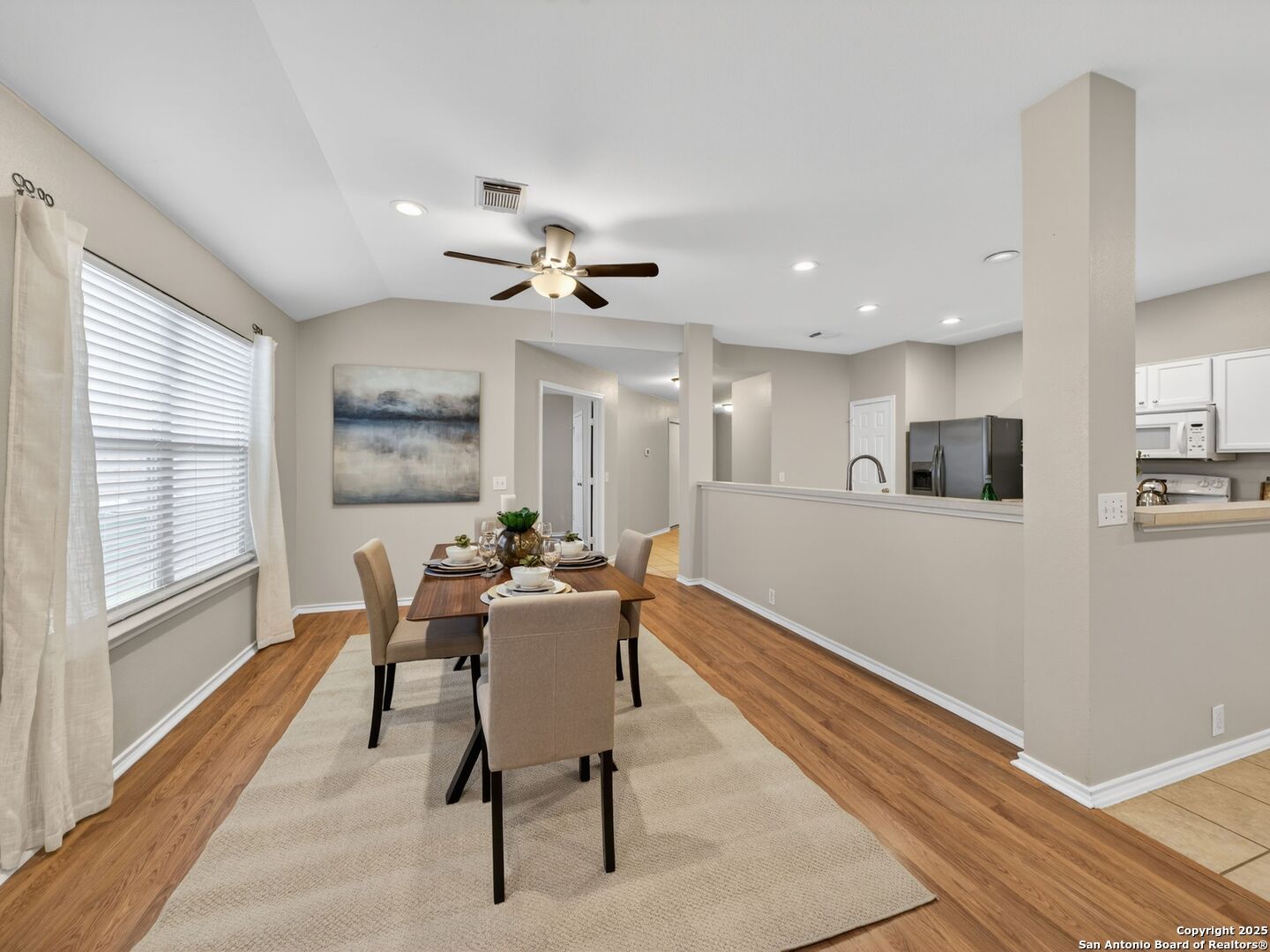 Dining area with a set table, cream rug, wide window, and open kitchen.