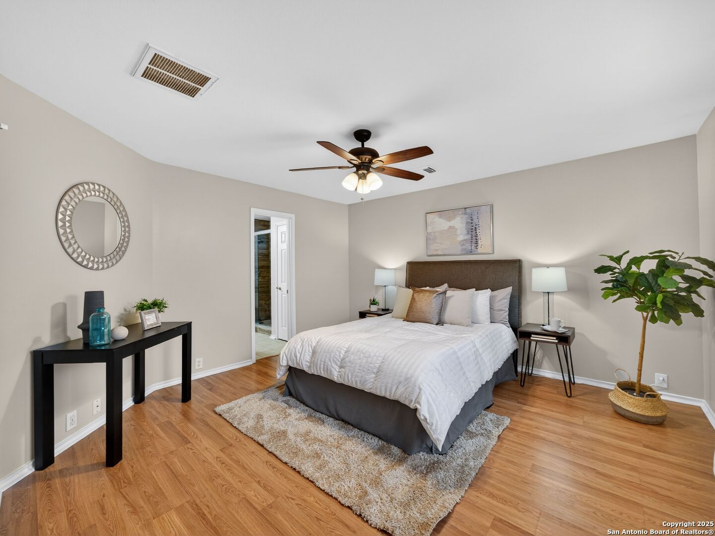 Furnished bedroom with a white-covered bed, ceiling fan, hardwood floors, and a potted plant.