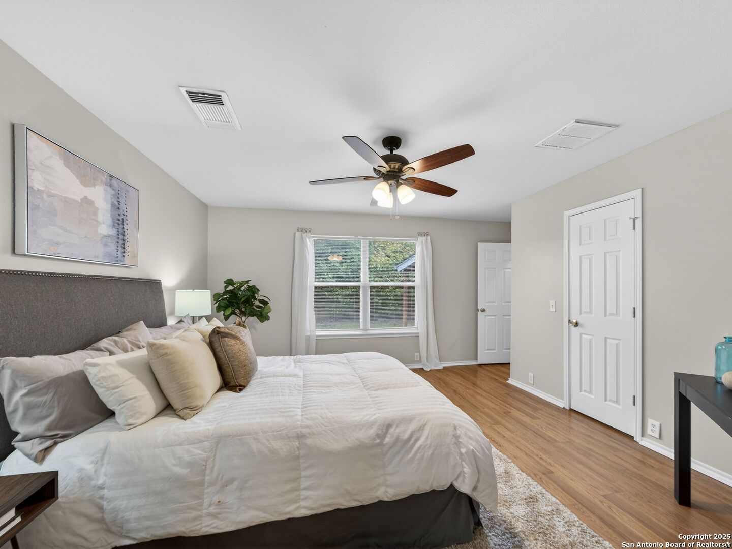 Furnished bedroom with a white bed, gray headboard, window overlooking greenery, and wood floors.