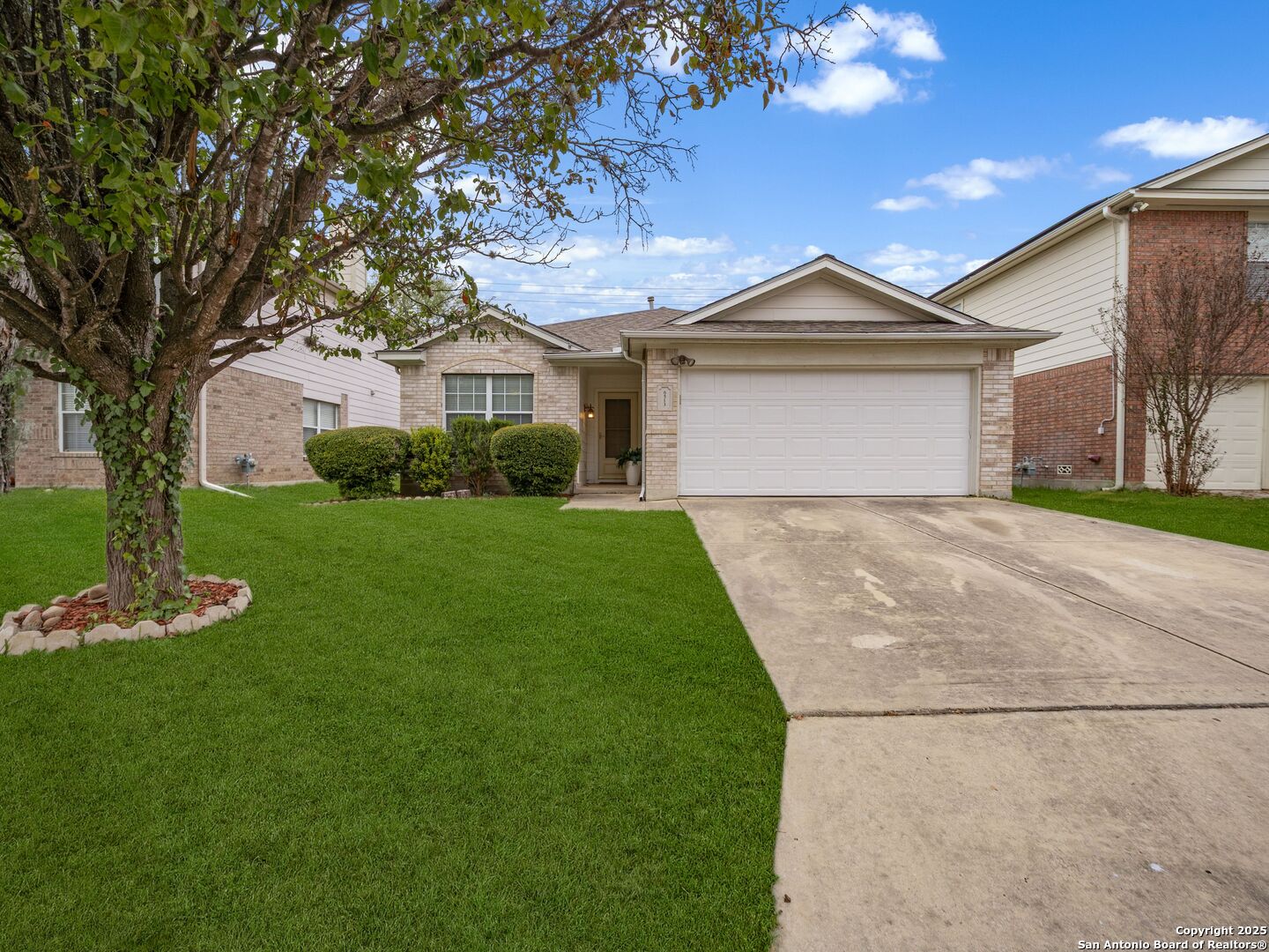 Single-story brick home with a two-car garage, broad driveway, green lawn, and mature tree.