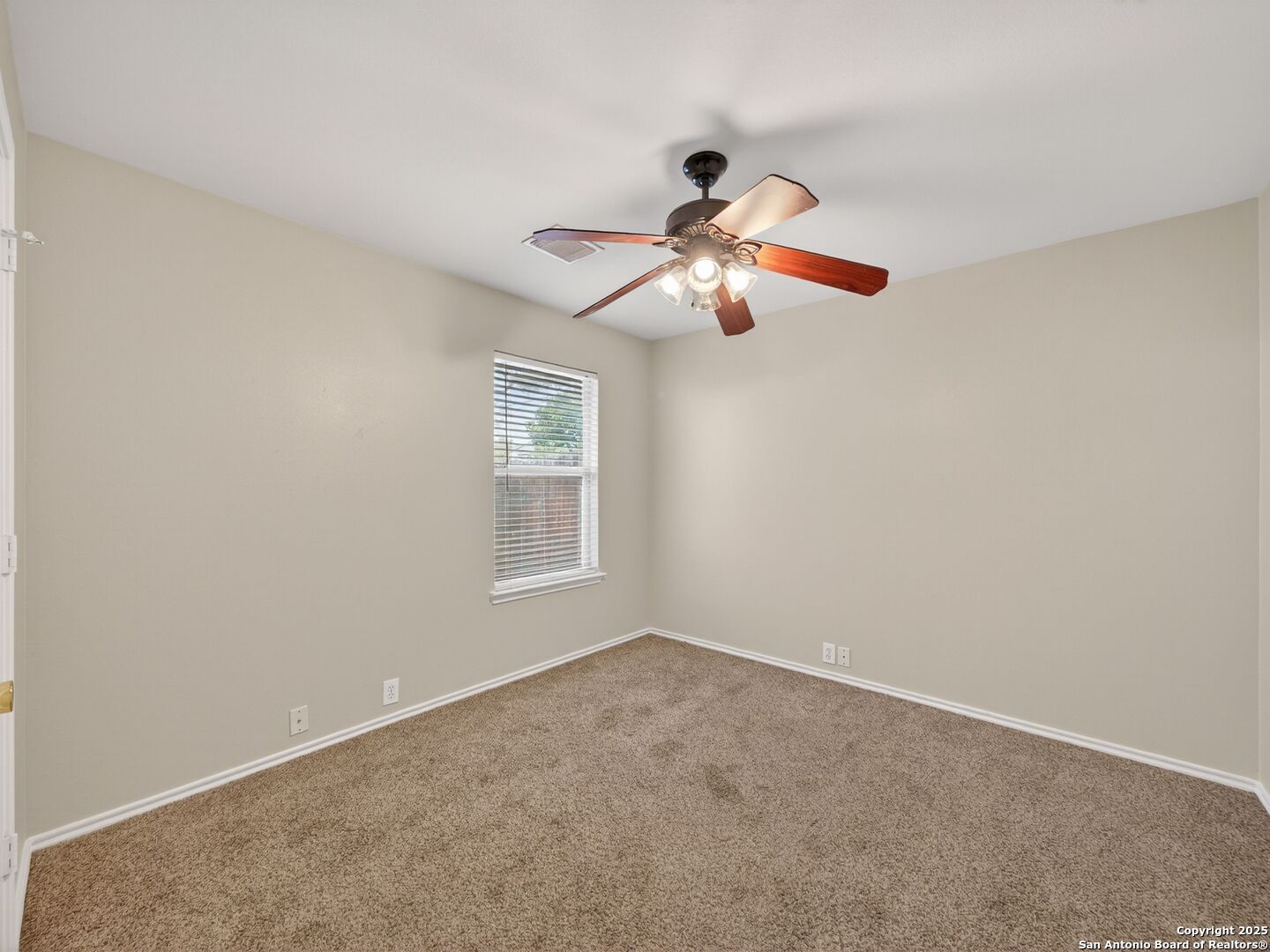Empty bedroom with tan carpet, a window with blinds, and a lit ceiling fan.