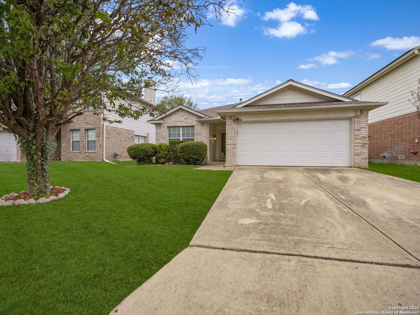 Single-story brick home with an attached two-car garage, broad driveway, and mature tree by the lawn.