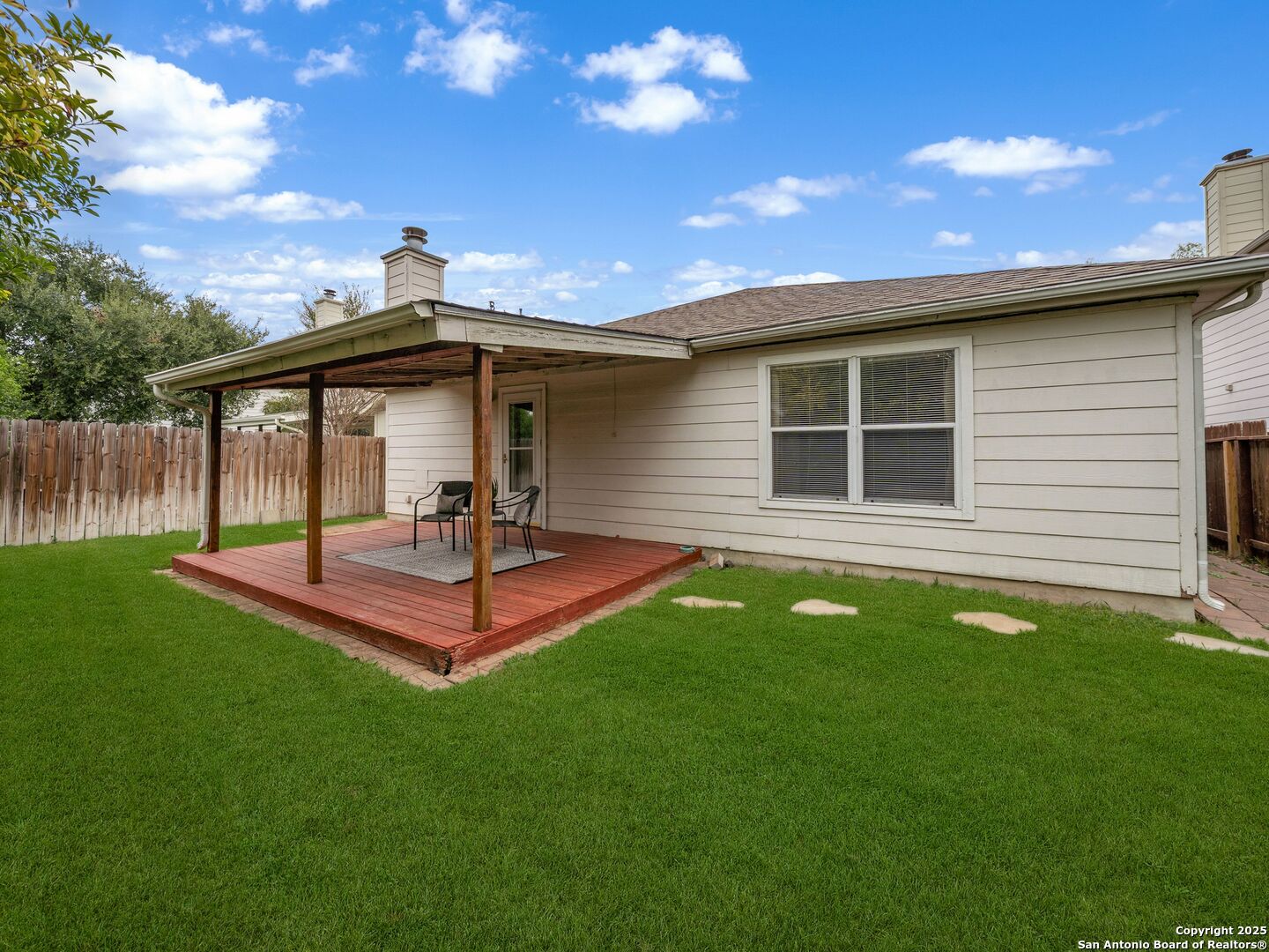 Rear of a light-colored home with a covered redwood deck, patio chairs, and a fenced lawn.