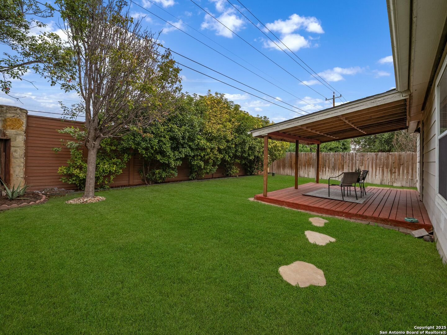 Fenced backyard with a covered wooden deck, outdoor seating, mature landscaping, and a broad green lawn.