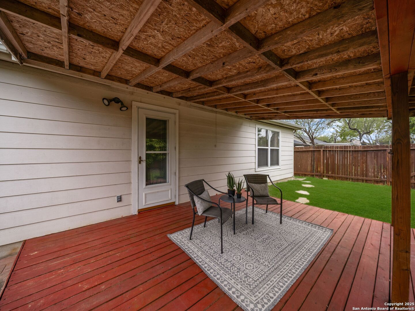 Covered backyard patio with exposed wooden beams, red deck boards, outdoor chairs, and a fenced lawn.