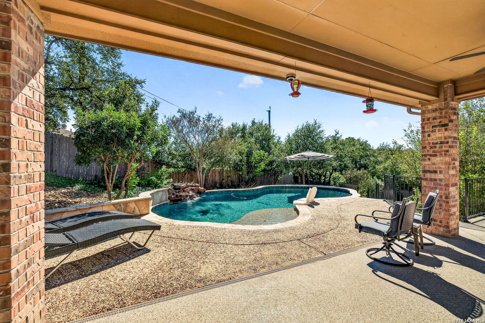 Covered patio overlooking a freeform swimming pool, with lounge chairs and a fenced, leafy yard.