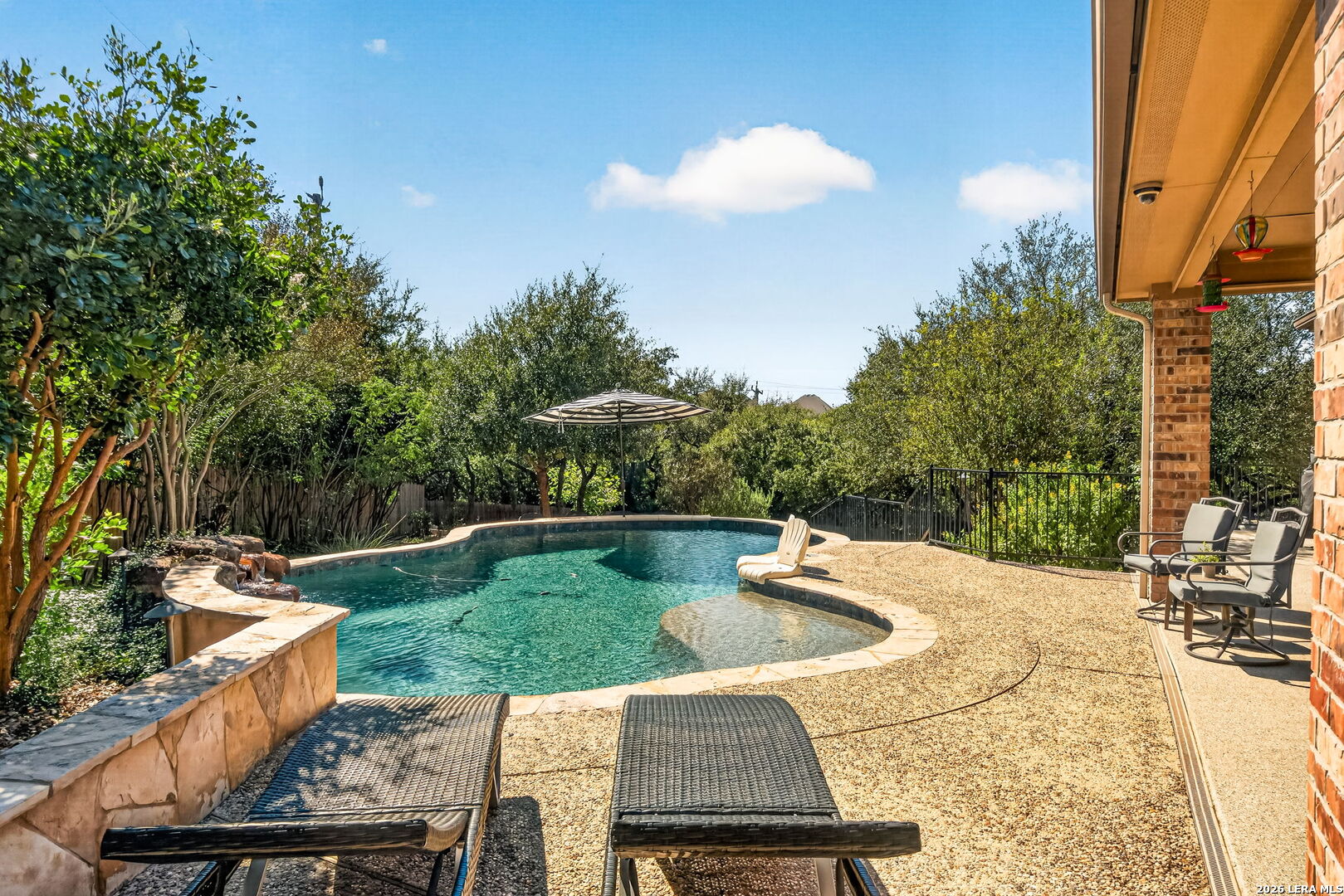 Freeform backyard pool with a shallow ledge, stone coping, patio chairs, and mature landscaping.