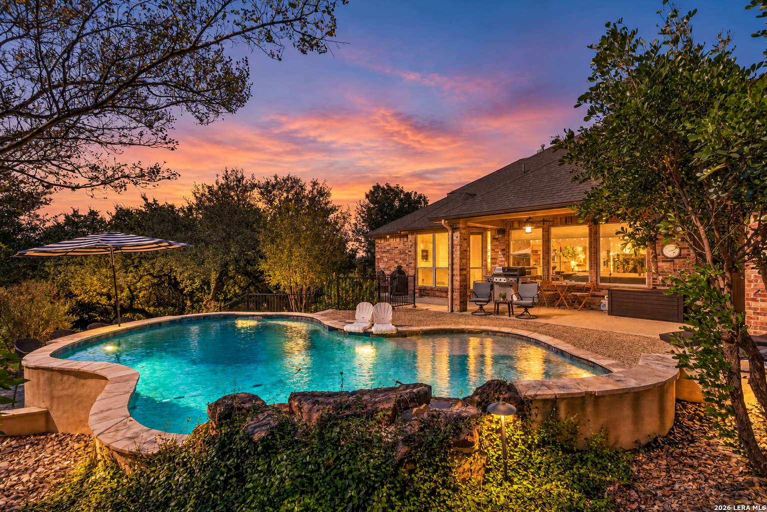 Backyard pool and brick patio at dusk, with outdoor seating and illuminated landscaping.