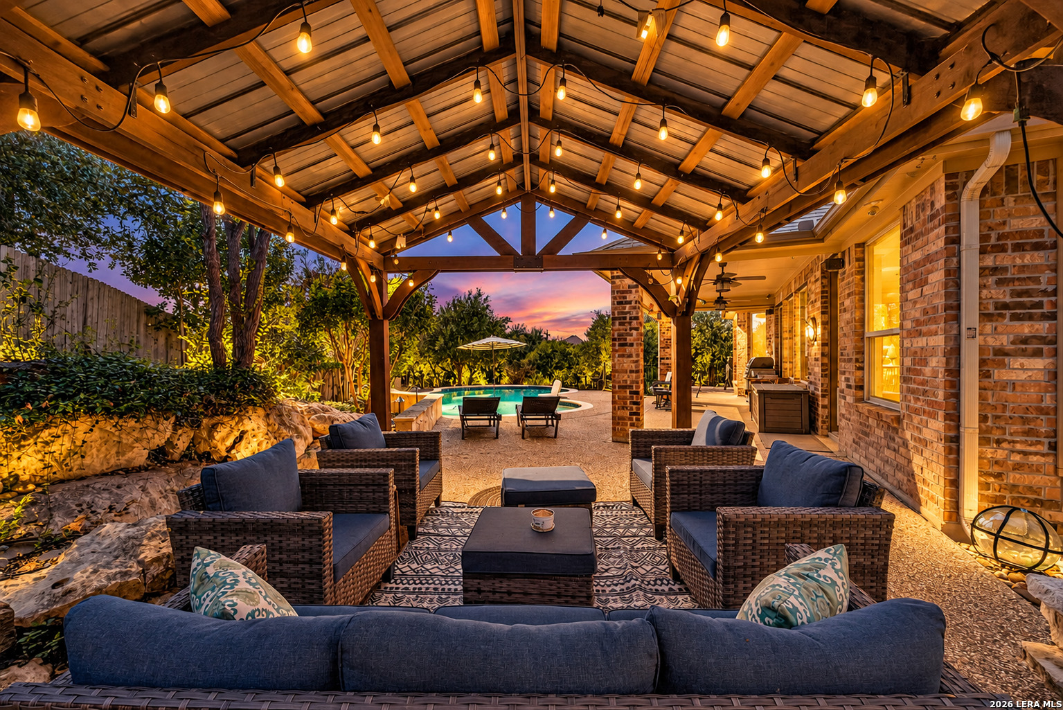 Covered patio with wicker seating, string lights, and a swimming pool at dusk