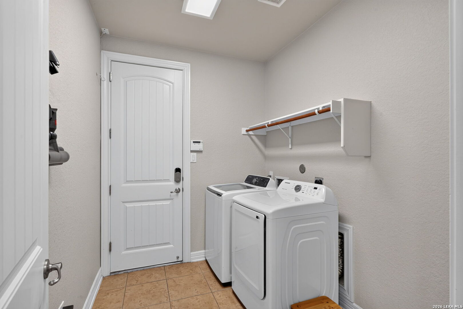 Laundry room with washer and dryer, wall-mounted shelf and hanging rod, and tan tile floor.